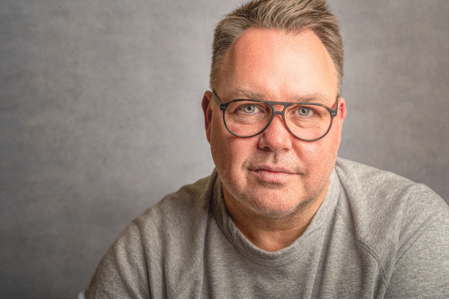 Professional headshot of man wearing glasses and grey sweater against neutral background.