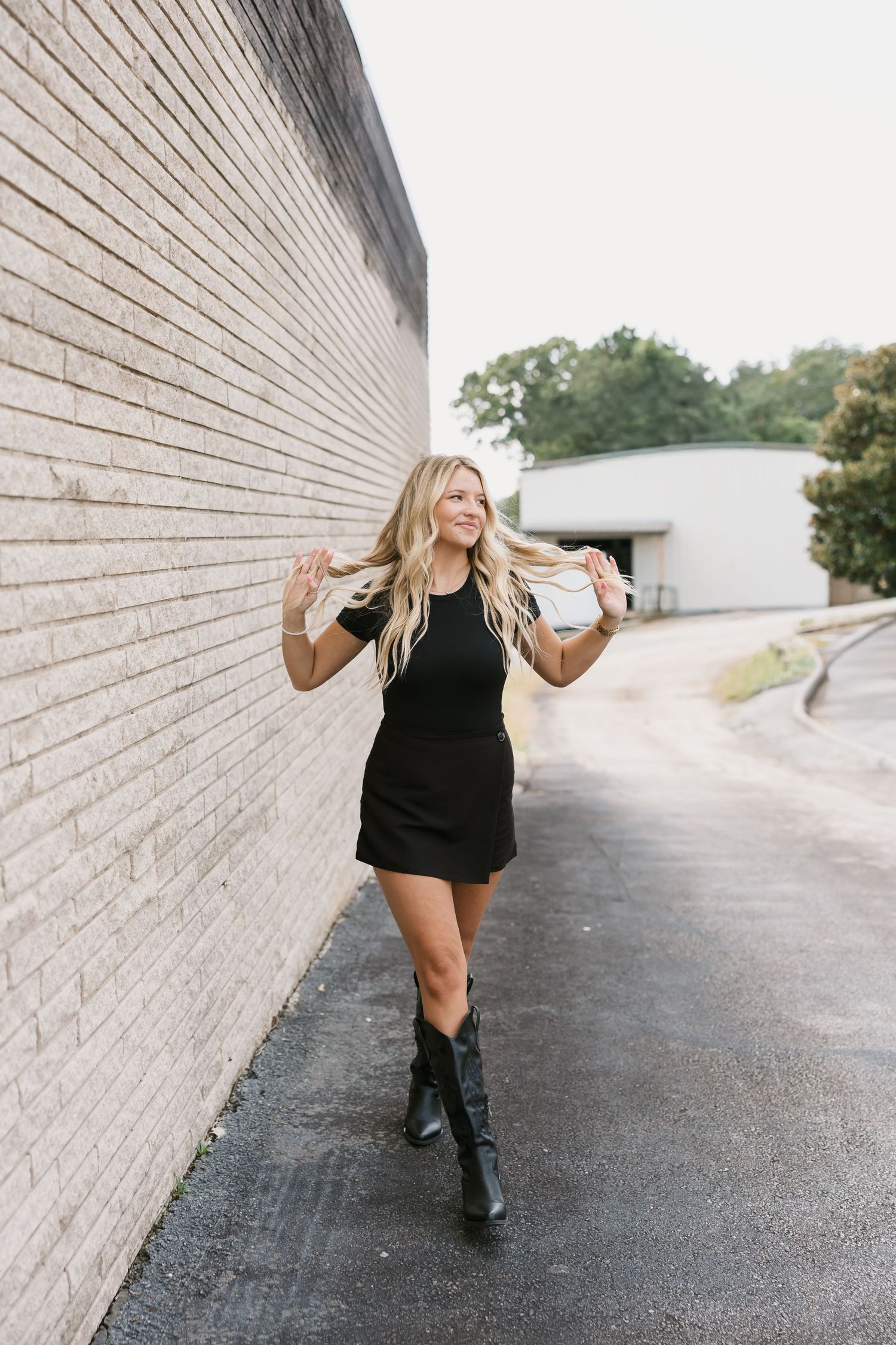 A figure in a black dress dances joyfully along a white brick wall and paved pathway in an urban setting.