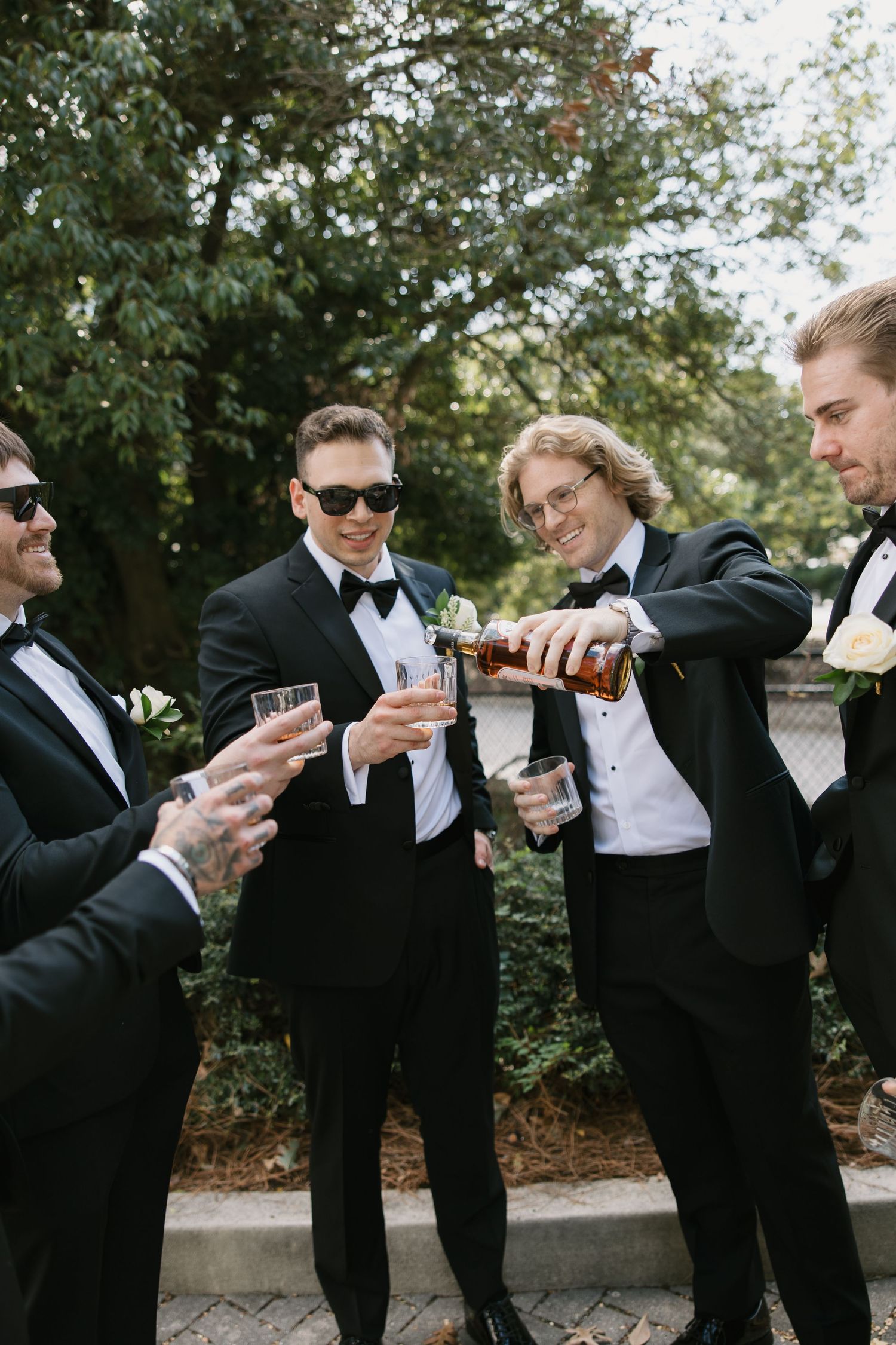 Groomsmen in black tuxedos and bow ties pour champagne together during outdoor wedding celebration.