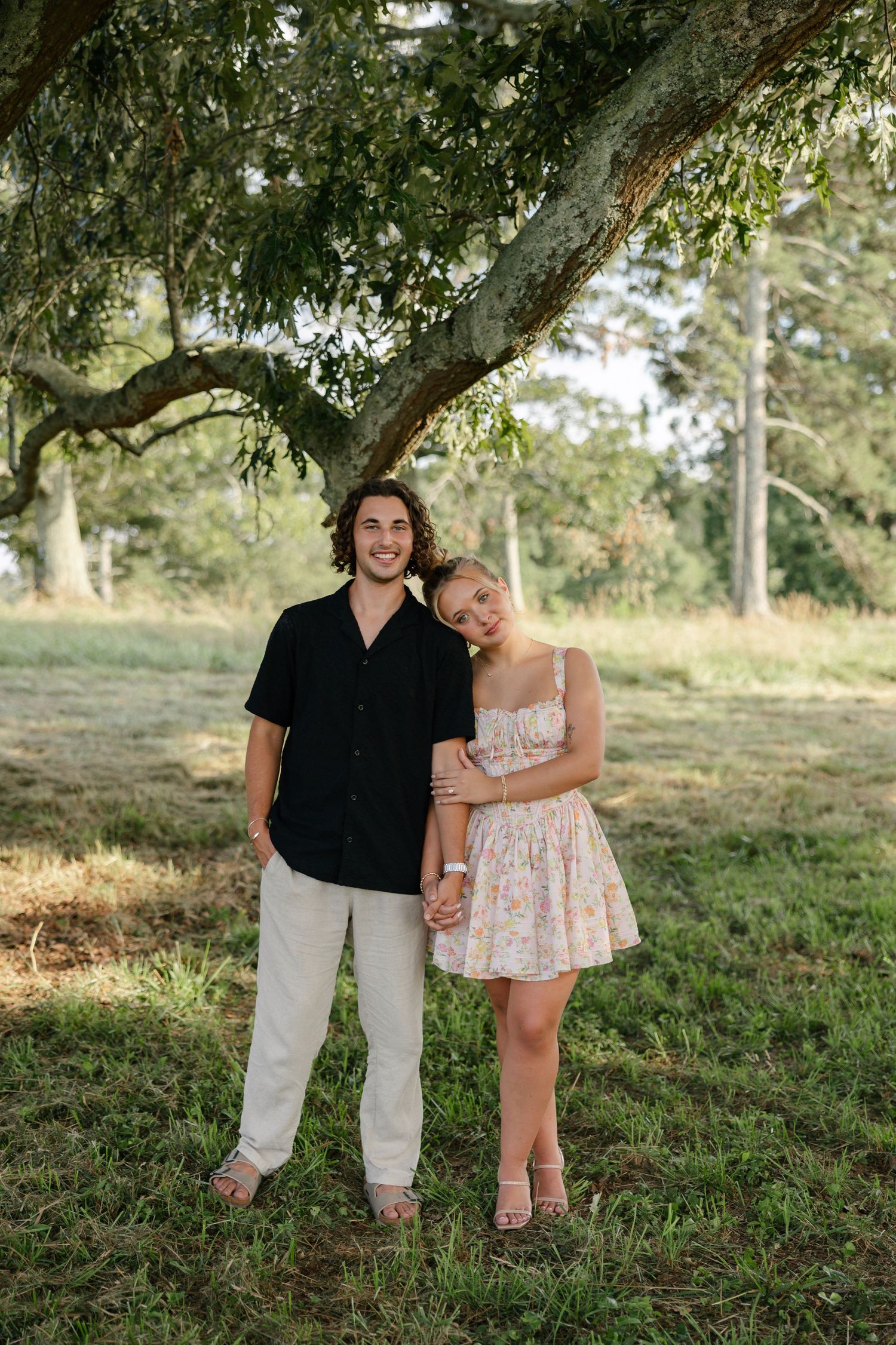 A couple embraces outdoors while sitting on a grassy hillside in golden sunlight during a summer photo shoot.