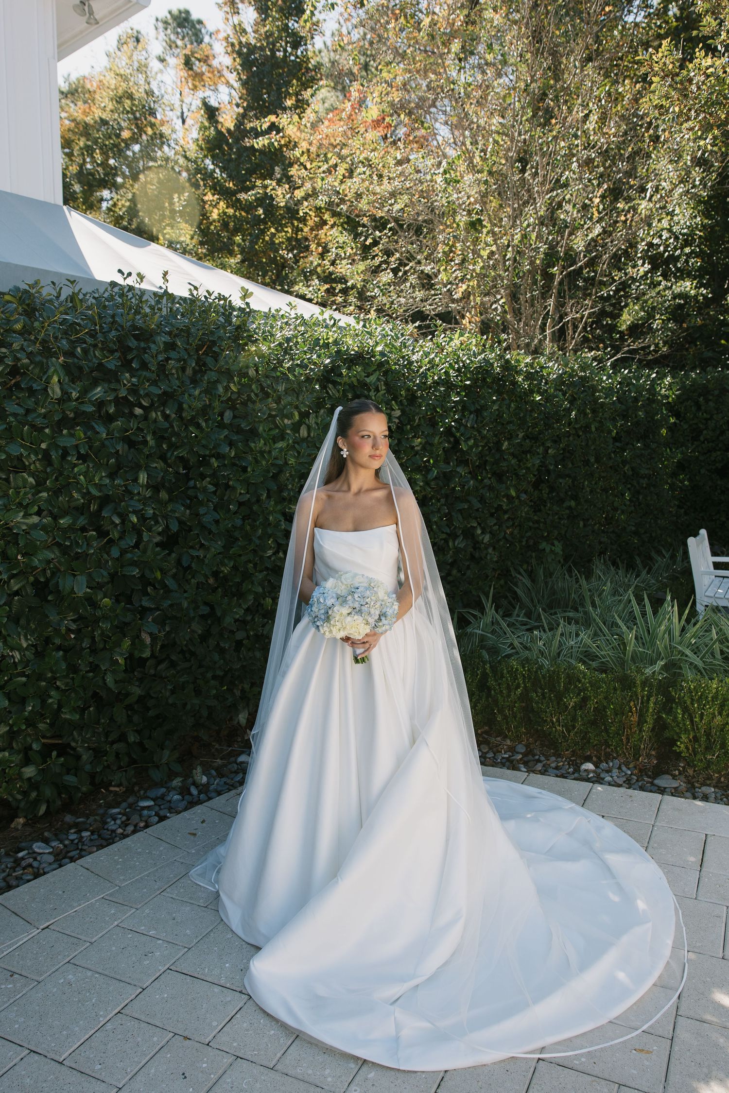 A bride in an elegant white satin ballgown with cathedral veil poses on stone pavers beside a lush garden hedge.