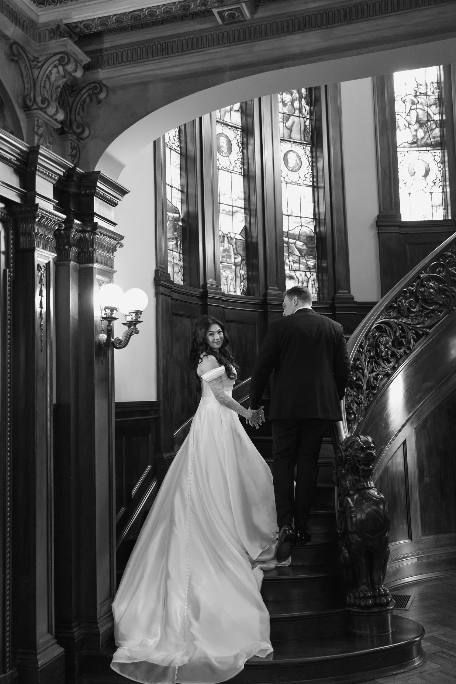 A bride in a flowing white ballgown poses gracefully on an elegant staircase in a historic mansion interior.