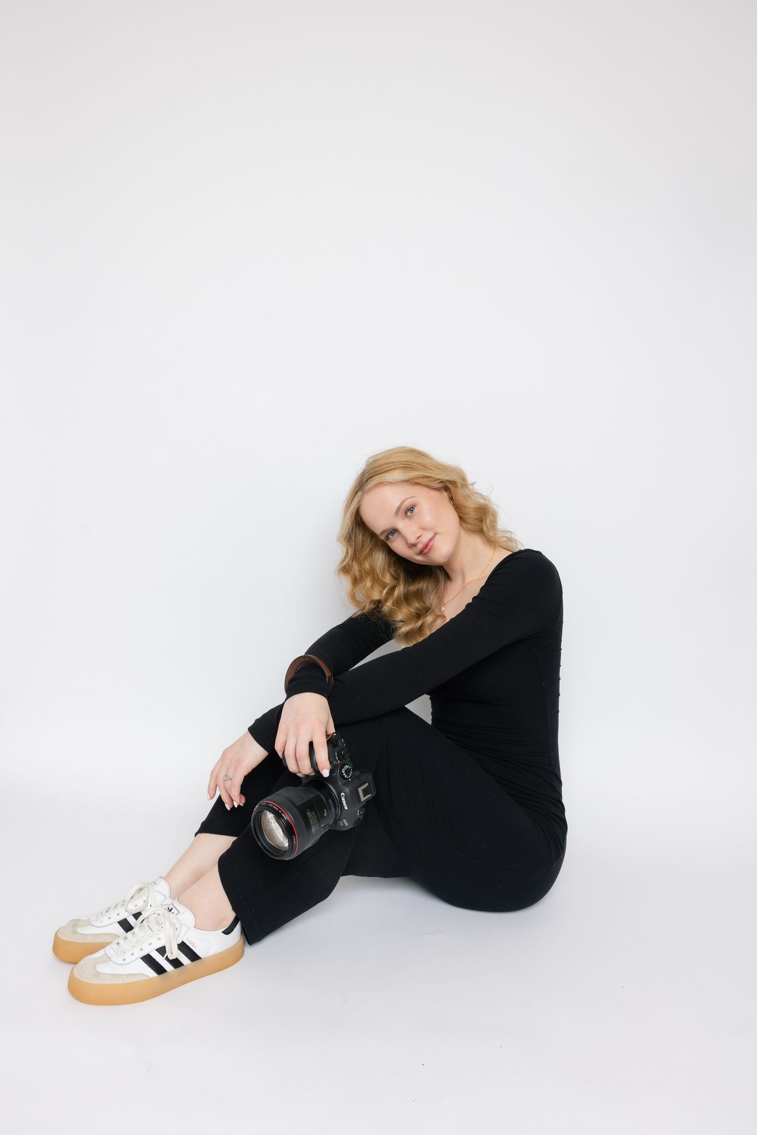 Woman in black outfit sitting on white floor wearing Adidas sneakers and looking away in minimalist setting.