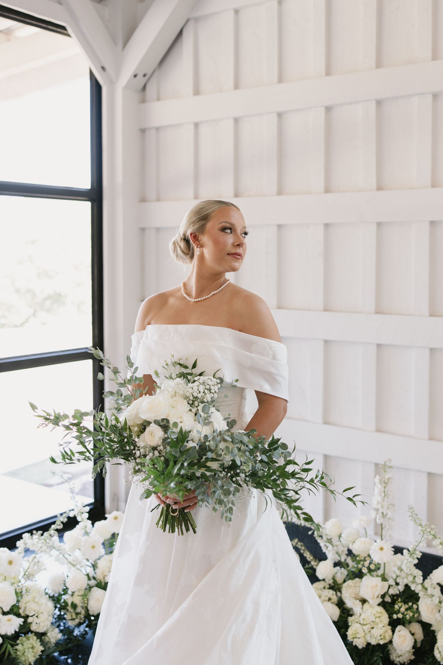 Elegant bride in off-shoulder white gown holds lush bouquet of white flowers and greenery in bright modern venue.