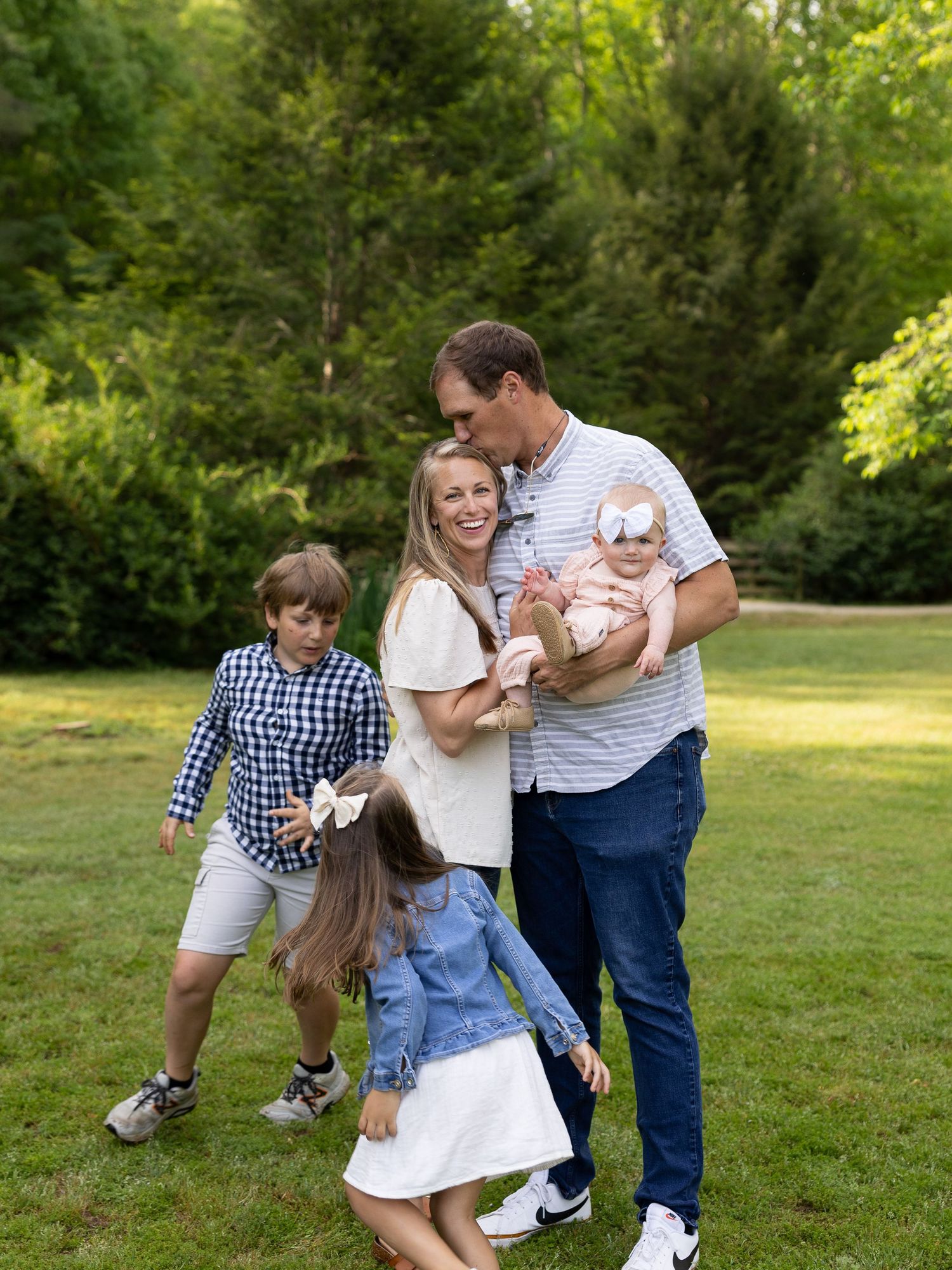 A joyful family of five poses outdoors on green grass, with tall trees in the background on a sunny day.