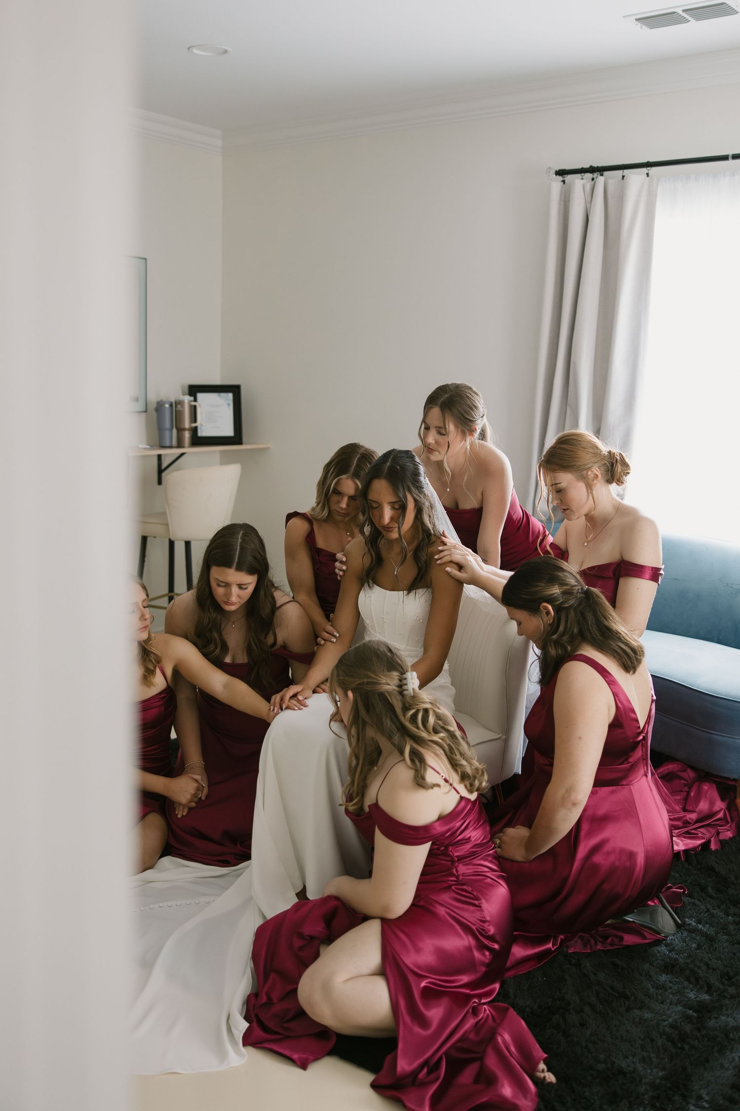 A group in burgundy bridesmaid dresses gather around someone in white on a dark carpet during wedding preparations.