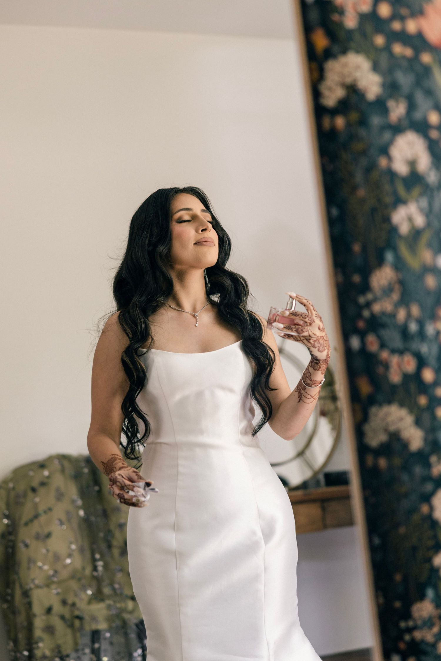 Simple elegant white fitted dress against floral wallpaper background with perfume bottle in an intimate setting.