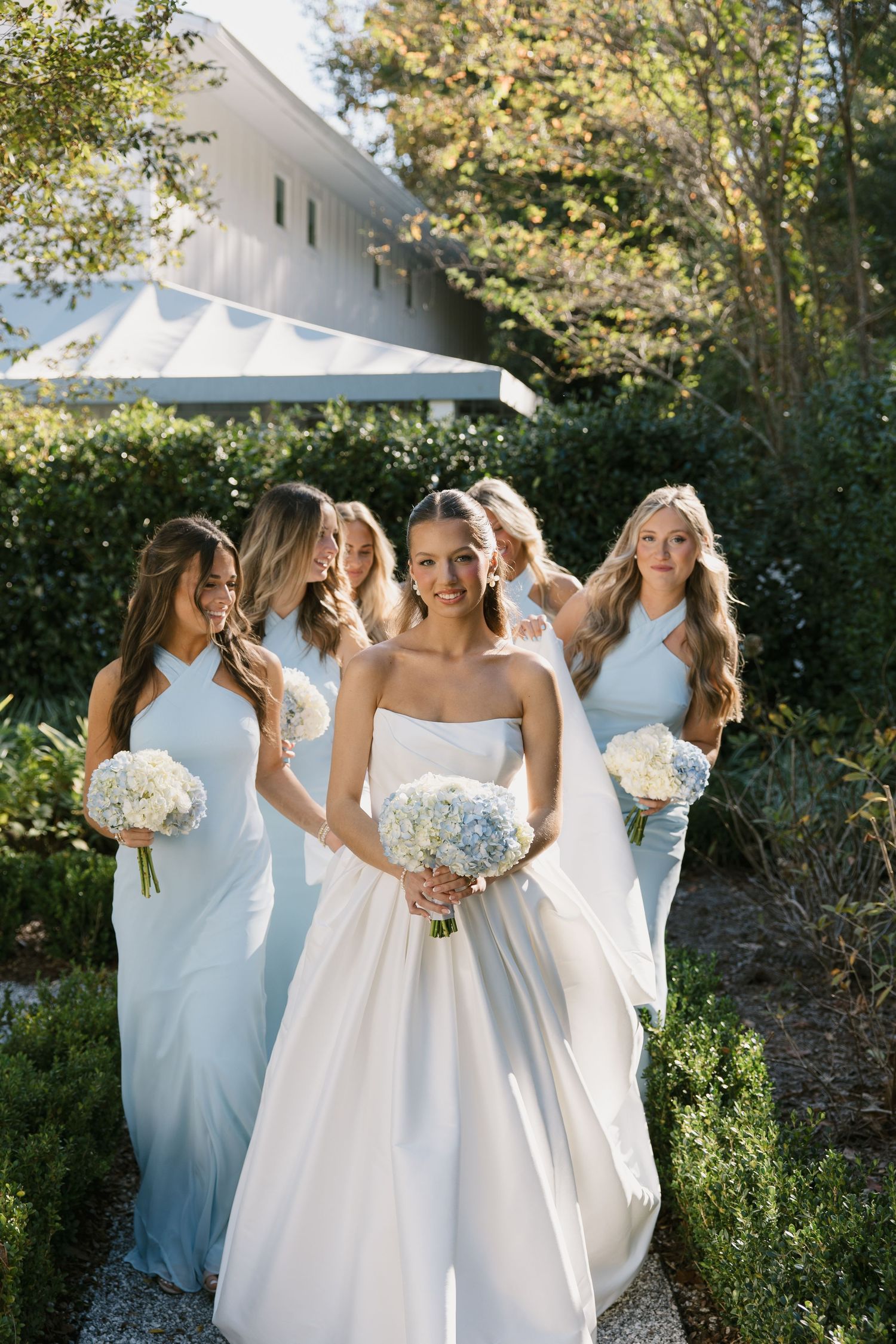 Bridesmaids in flowing white dresses hold bouquets while standing together in a sunlit garden setting.