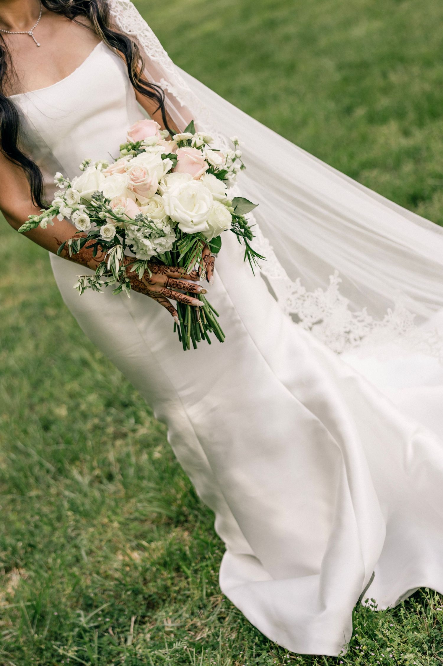 A close-up of an elegant white wedding dress with a flowing train and beautiful white and green floral bouquet on grass.