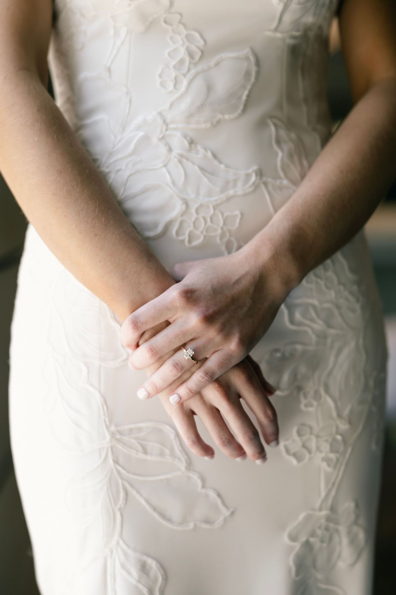 Close-up of hands wearing engagement ring on white lace wedding dress with floral embroidery pattern.