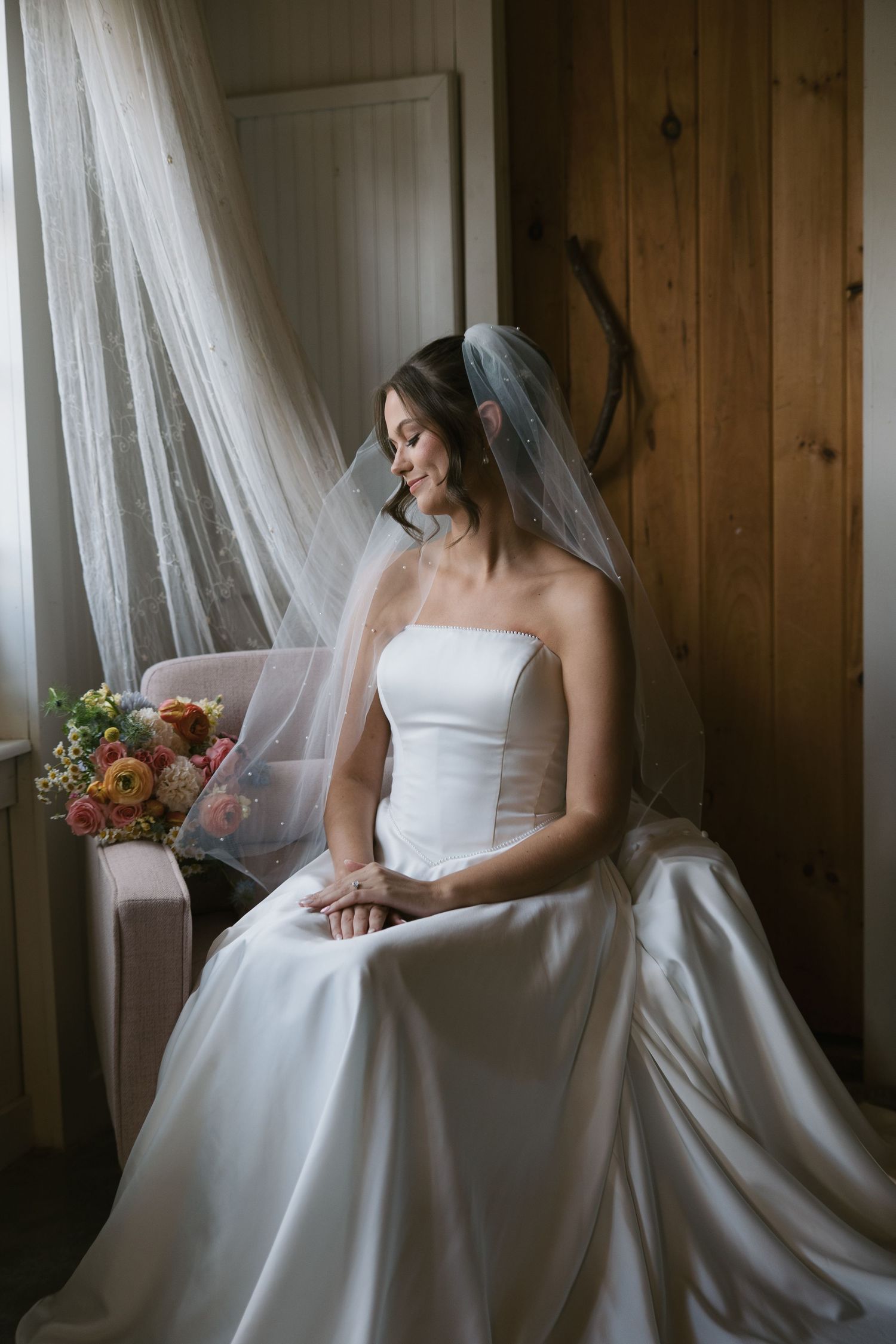 Elegant bride in flowing white gown sits by window as light filters through sheer curtains in rustic wooden room.