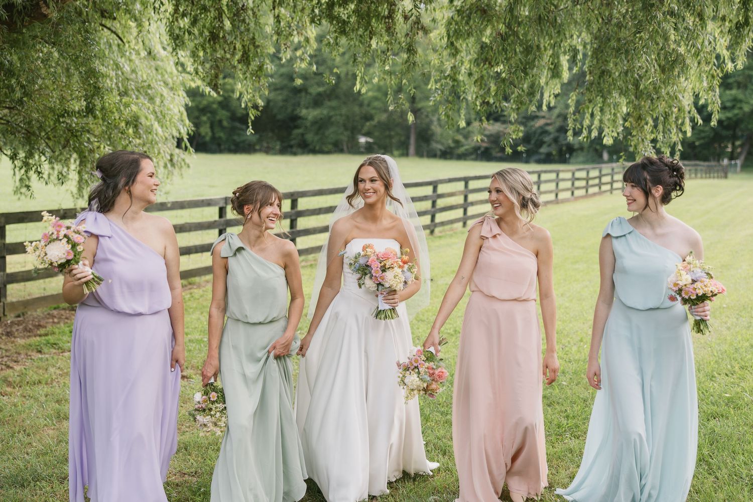 Bridesmaids in pastel pink, blue and lavender dresses gather together on a lush green lawn near wooden fences.