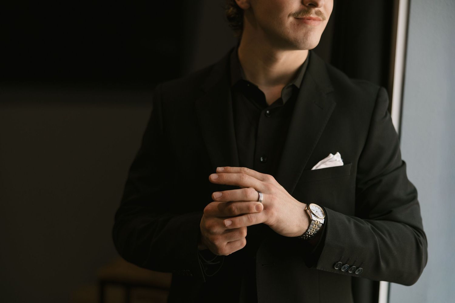 Professional in a black suit jacket and dress shirt adjusts his ring while standing in moody lighting.