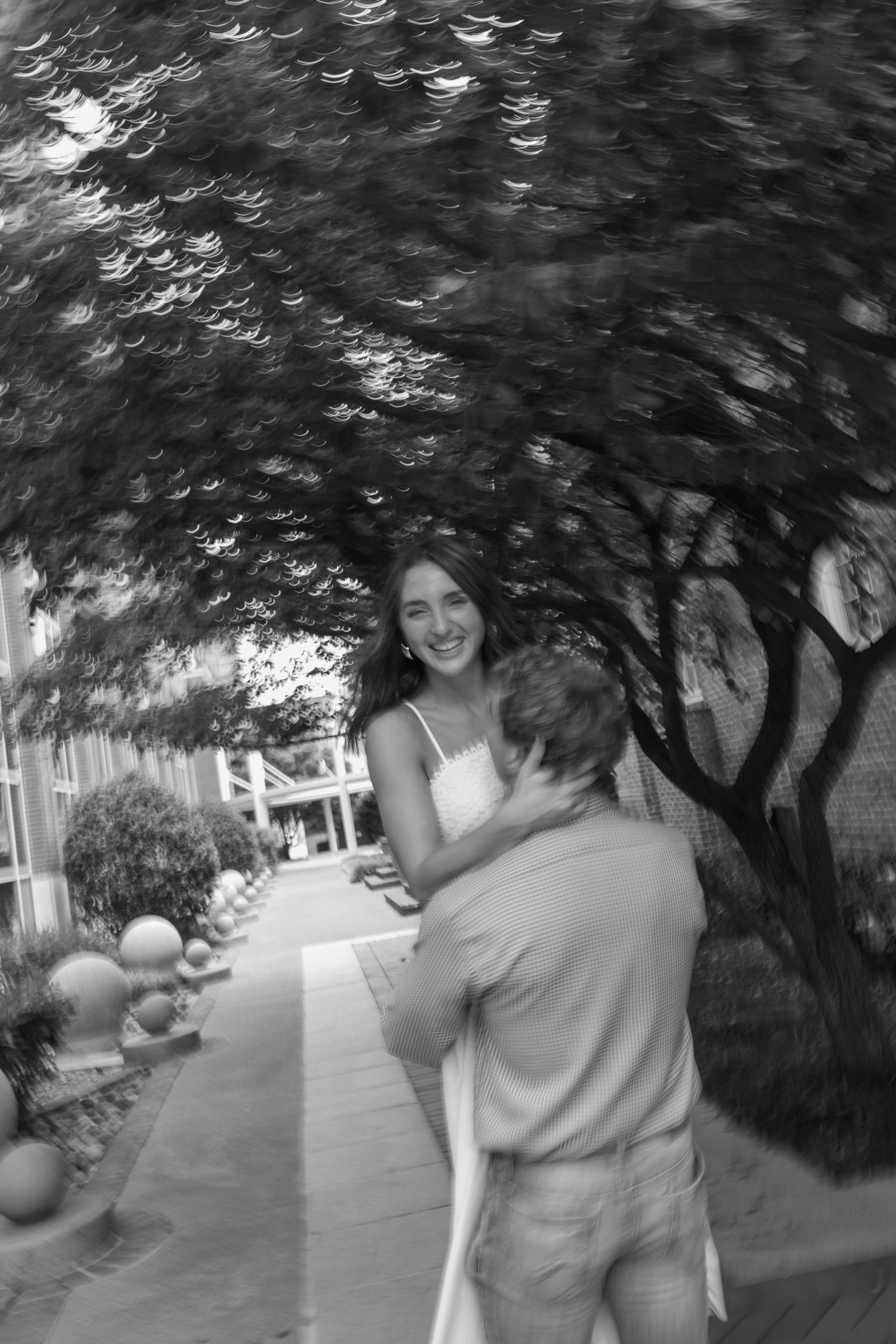 Black and white photo of two people embracing under a leafy tree canopy along a garden pathway.