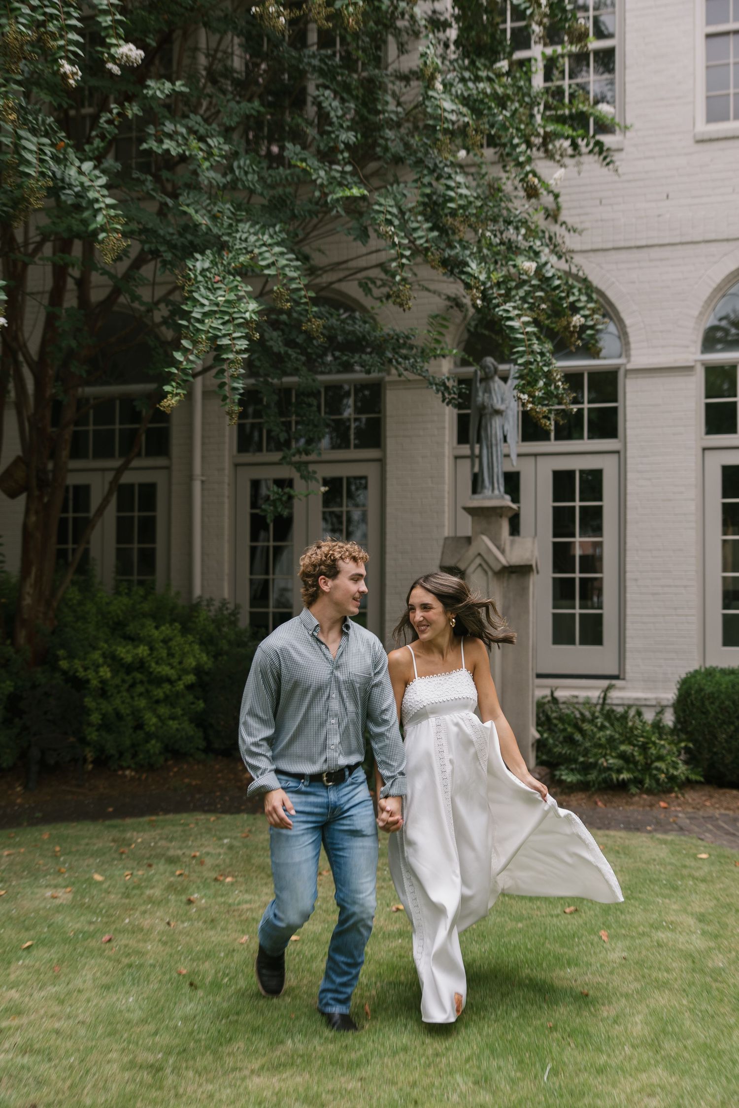 A couple in casual attire playfully runs through a green lawn in front of a light-colored building.