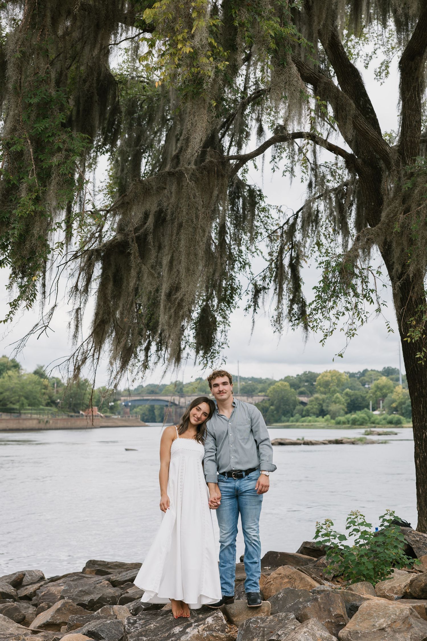 Couple enjoys romantic moment on rocky waterfront beneath moss-draped trees by tranquil lake at sunset.
