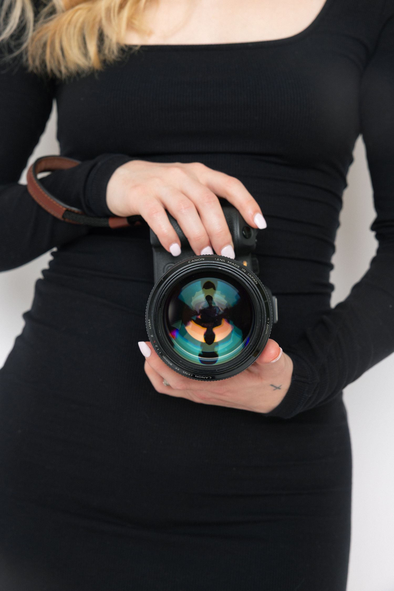 Person wearing black long-sleeve dress holds up a camera lens showing reflective glass elements against white background.