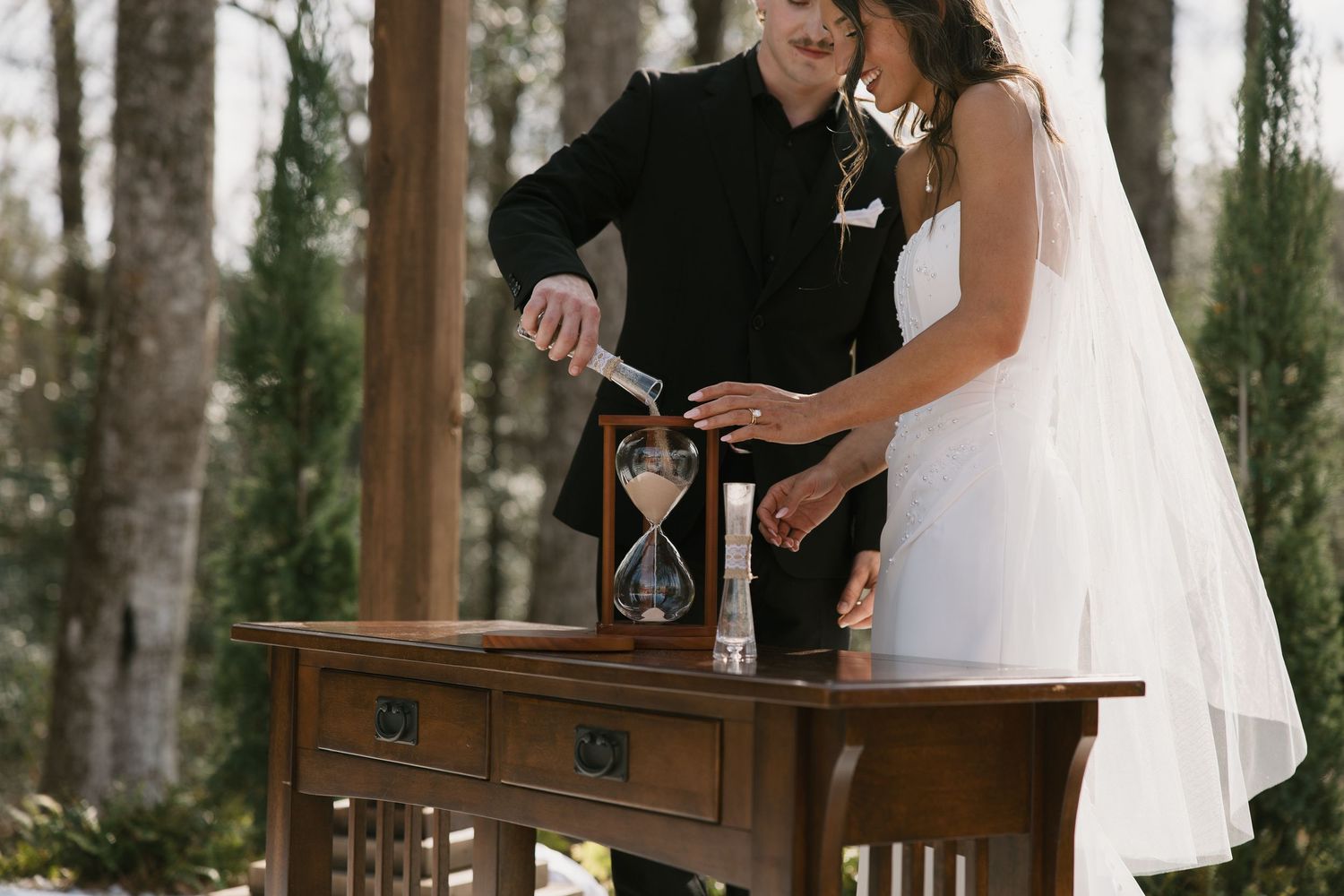 A couple pours sand into an hourglass during a unity ceremony at an outdoor wedding.