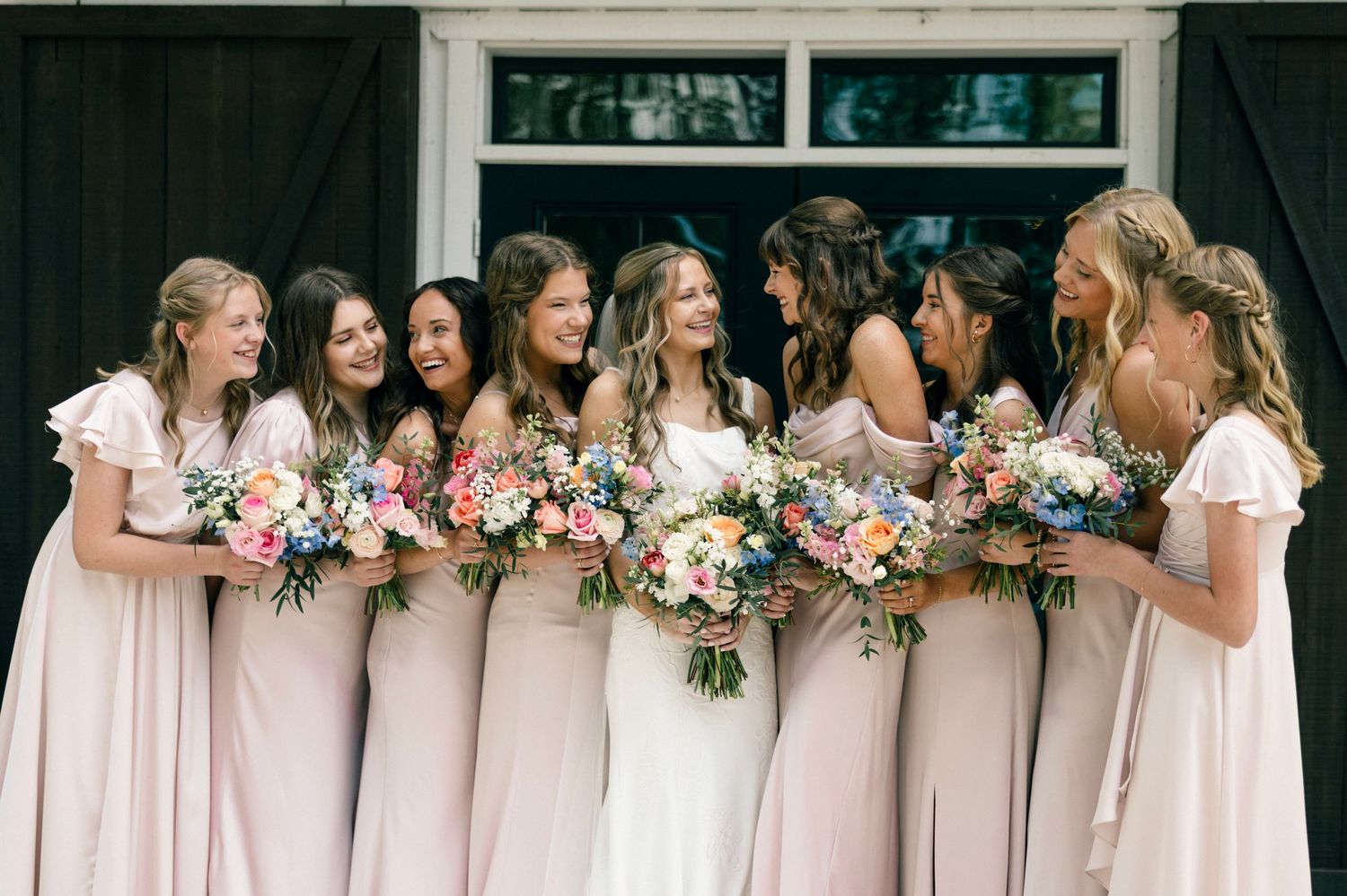 Bridesmaids in blush pink dresses share laughs while holding vibrant pink and white floral bouquets.