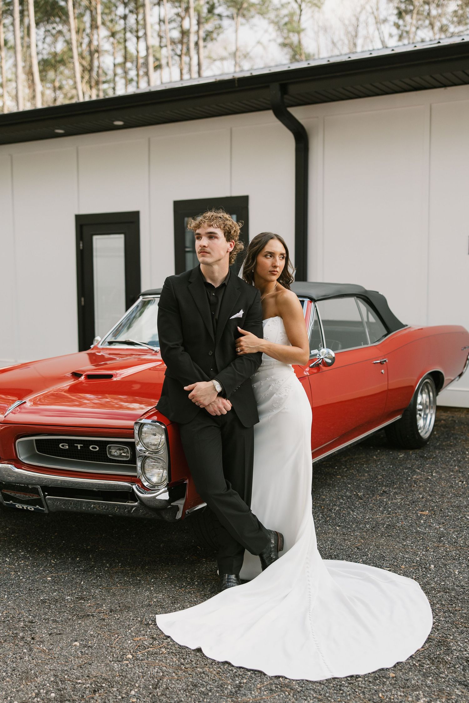 A couple poses for wedding photos next to a vintage red GTO car, with the bride wearing a flowing white gown.