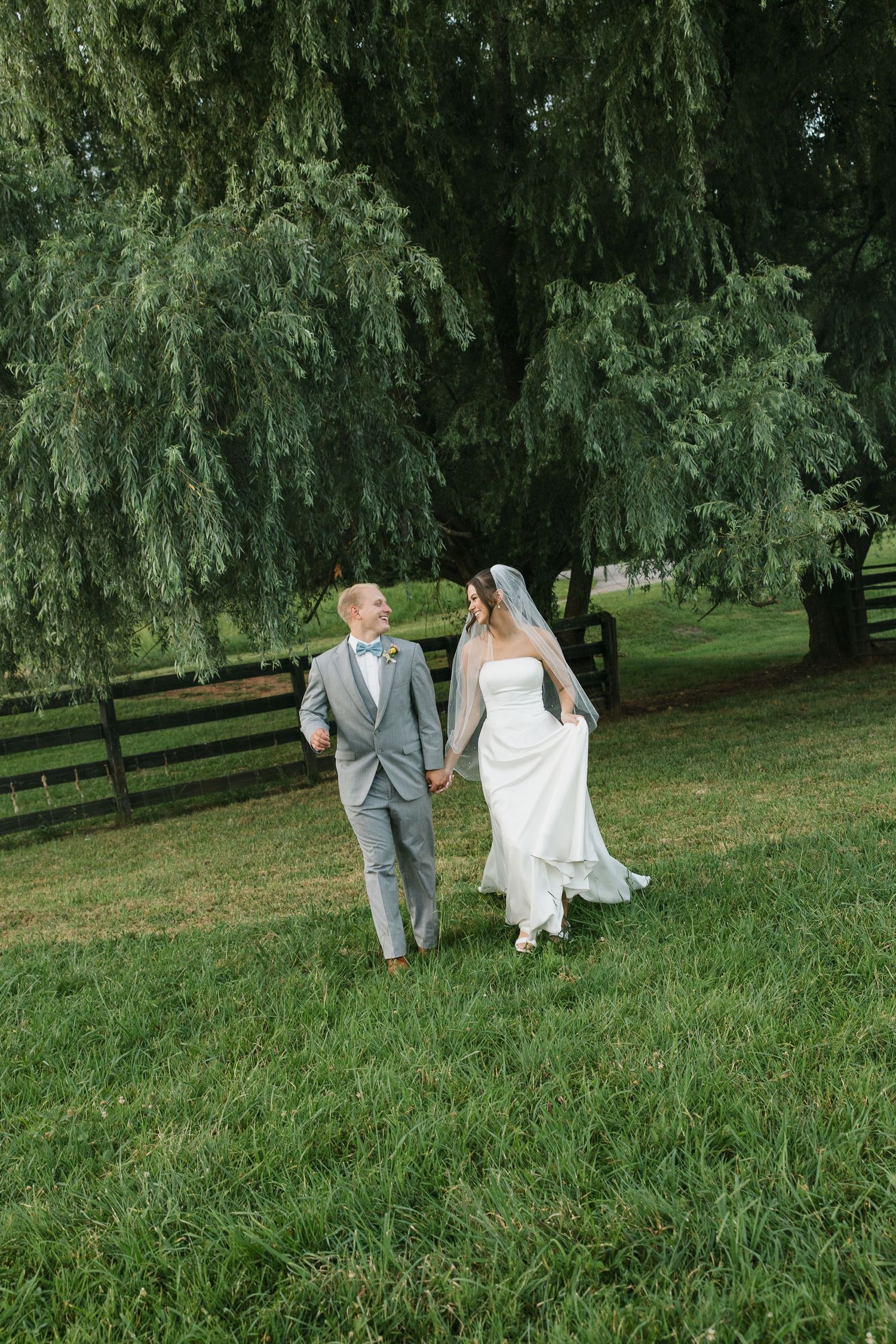 A couple in wedding attire walk together across a lush green lawn beneath drooping willow trees on a sunny day.