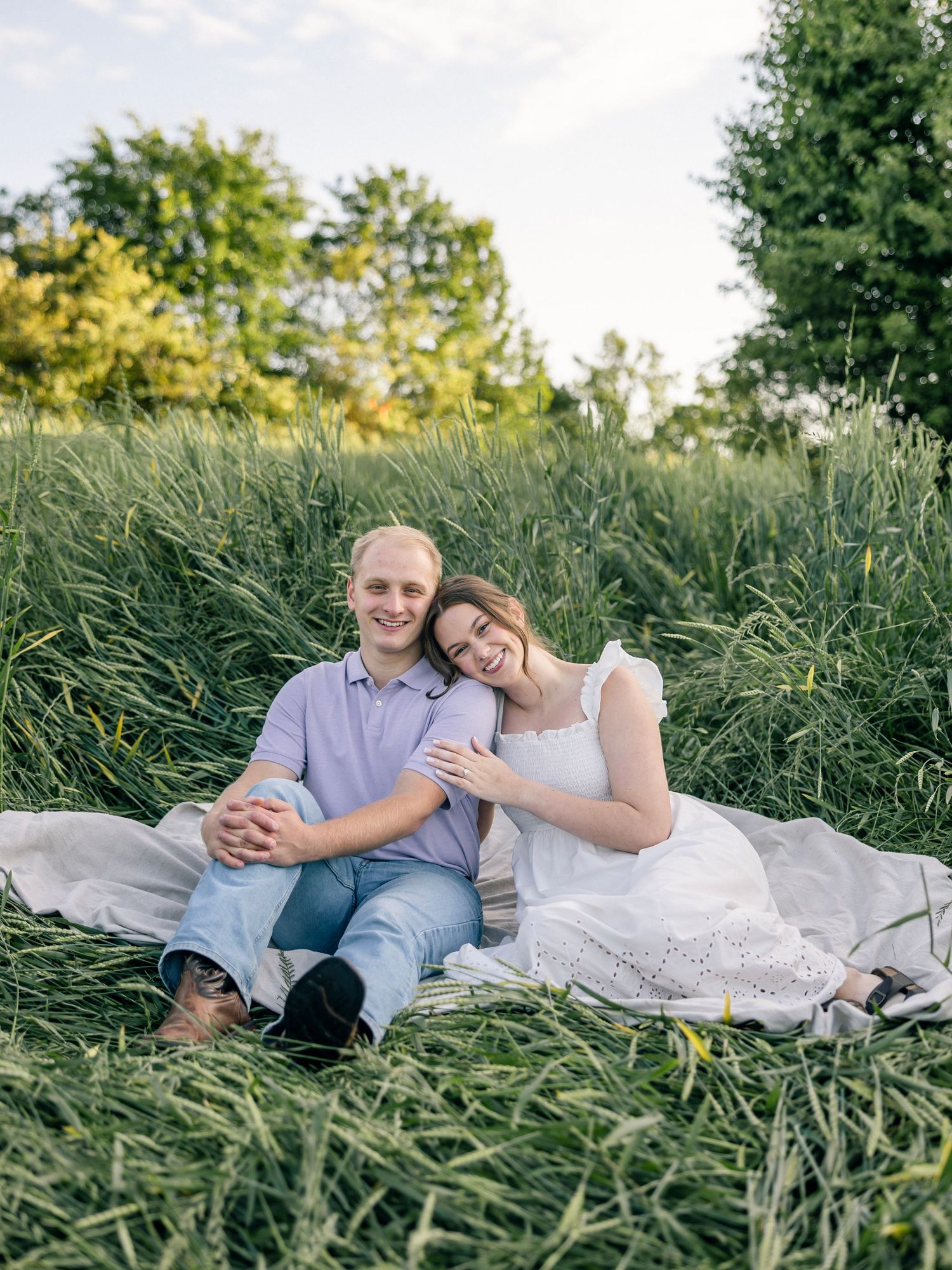 Couple sitting together on white blanket in tall grass field during golden hour.