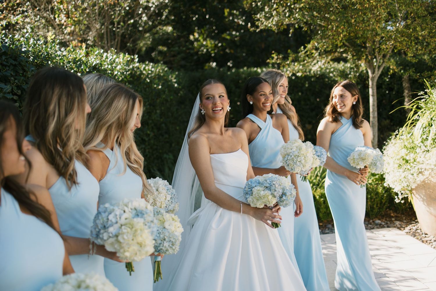 Bridesmaids in light blue dresses hold white and yellow bouquets while walking outdoors in a garden setting.