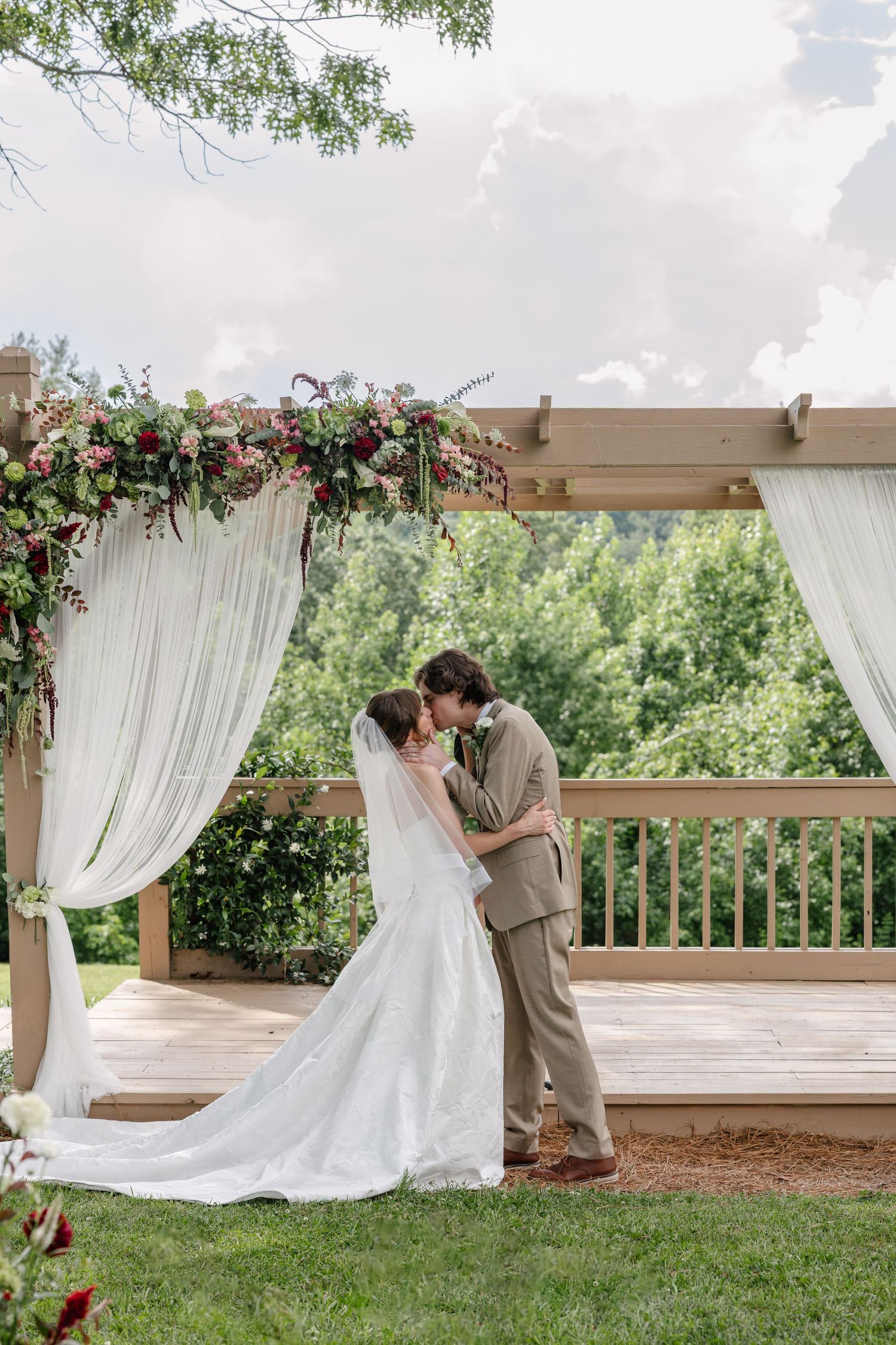 A couple embraces under an elegant draped wedding arch adorned with pink and white flowers on an outdoor wooden deck.