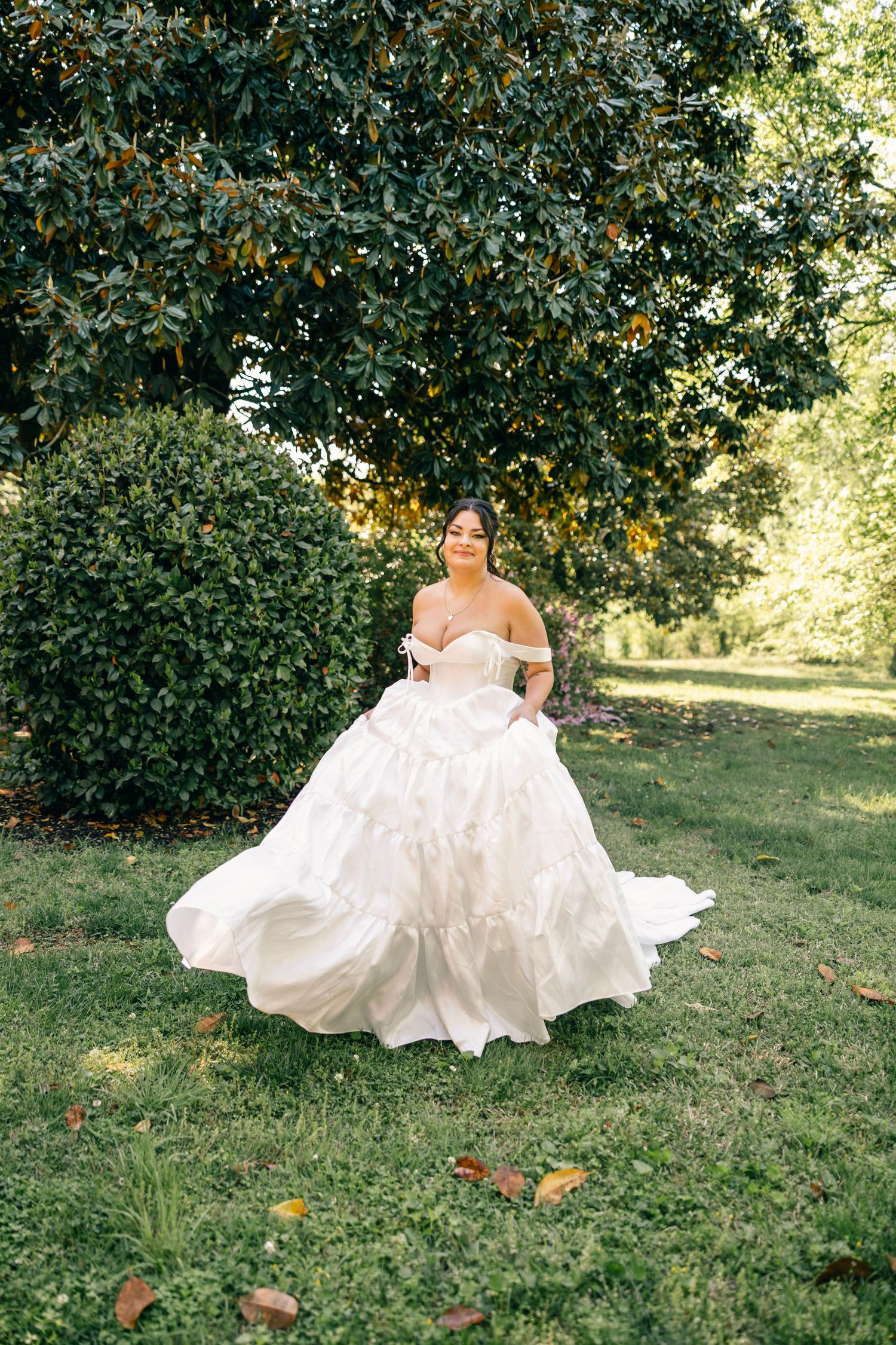 A bride in an elegant off-shoulder white ballgown stands on grass beneath a tree in a garden setting.