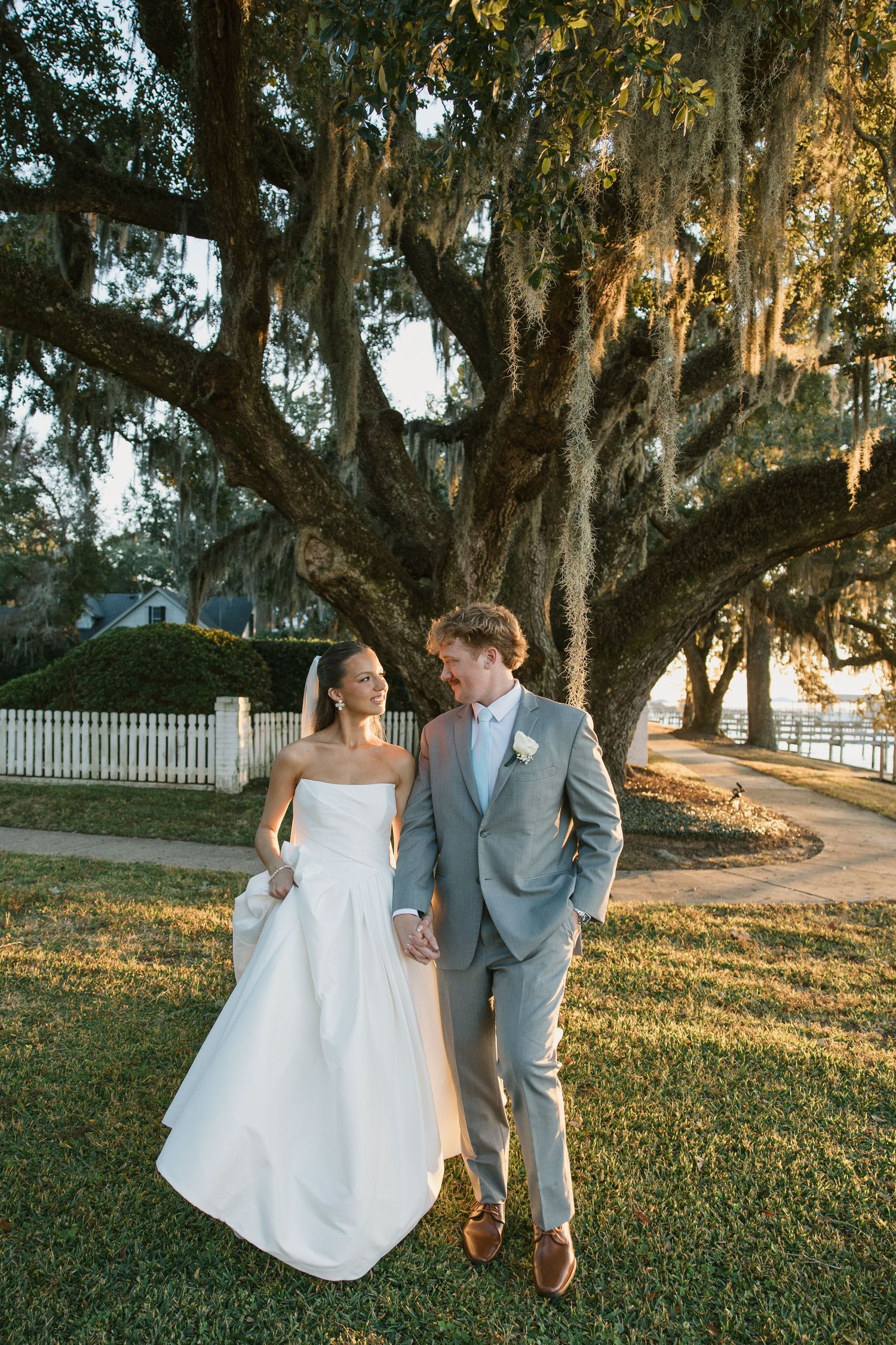 A couple in wedding attire walks together under Spanish moss-draped oak trees during golden hour.