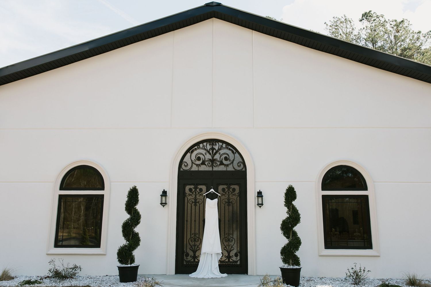 White wedding chapel entrance with symmetrical windows, spiral topiaries, and elegant black arched doorway with curtains.