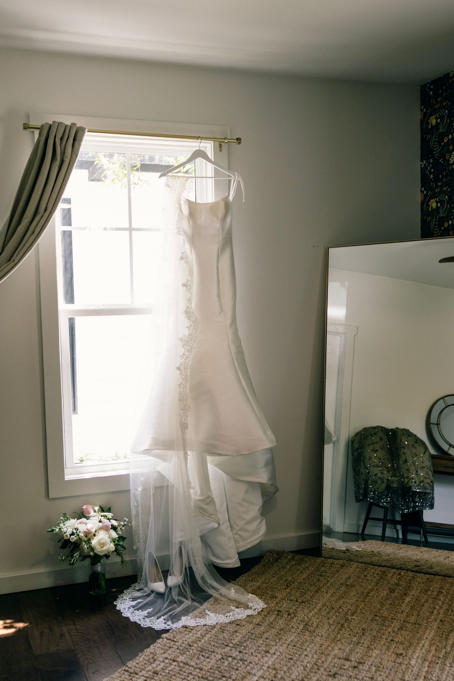 Elegant white wedding dress with lace and train hanging by window with natural light streaming in and flowers below.