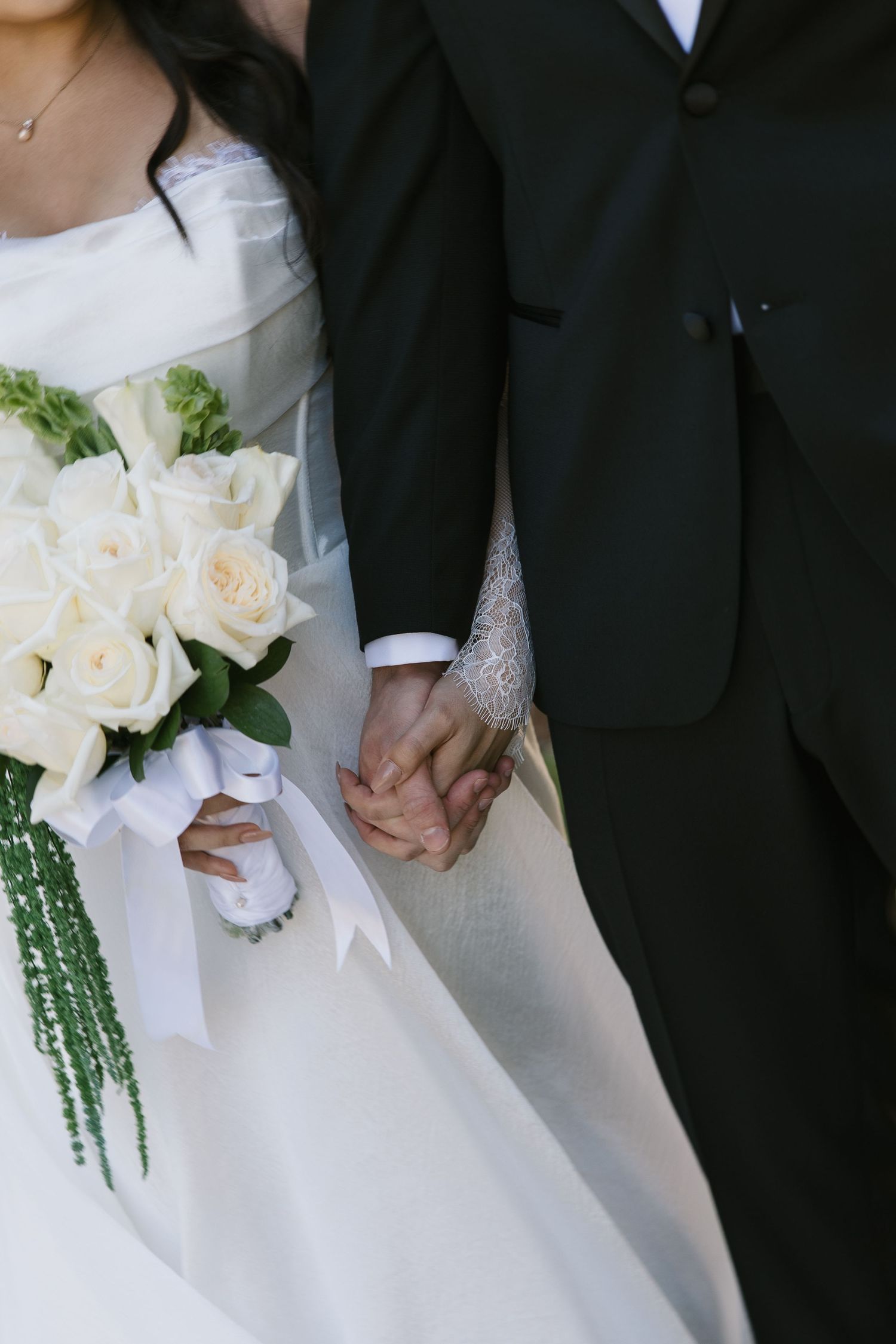 Close-up of newlywed couple holding hands with white roses and greenery bouquet against black and white attire.