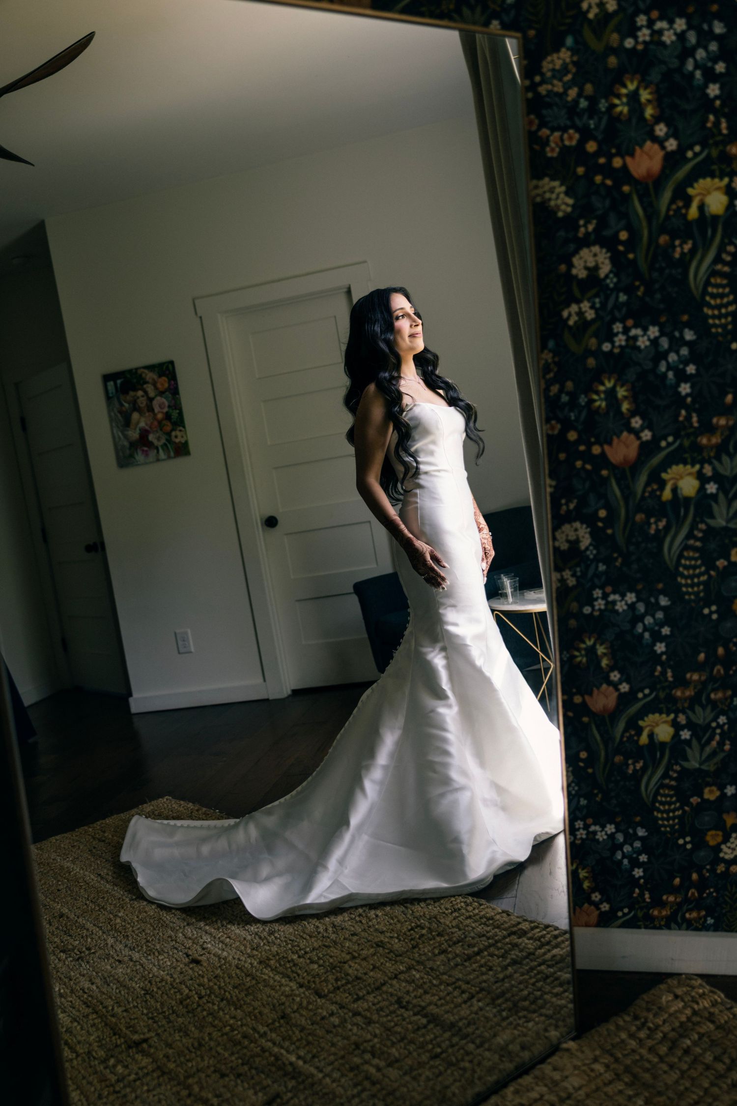A stunning mermaid-style wedding gown with a long train is showcased in a room with floral wallpaper.