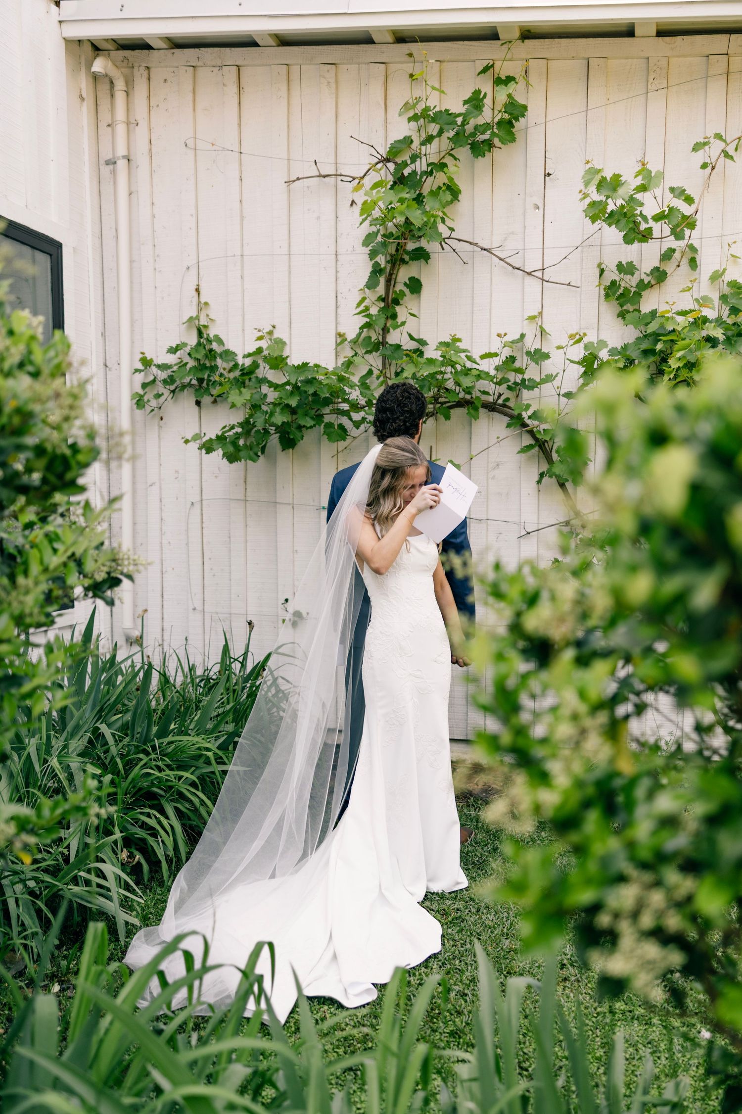 A couple in wedding attire embrace by a rustic white barn wall with climbing vines and garden greenery.
