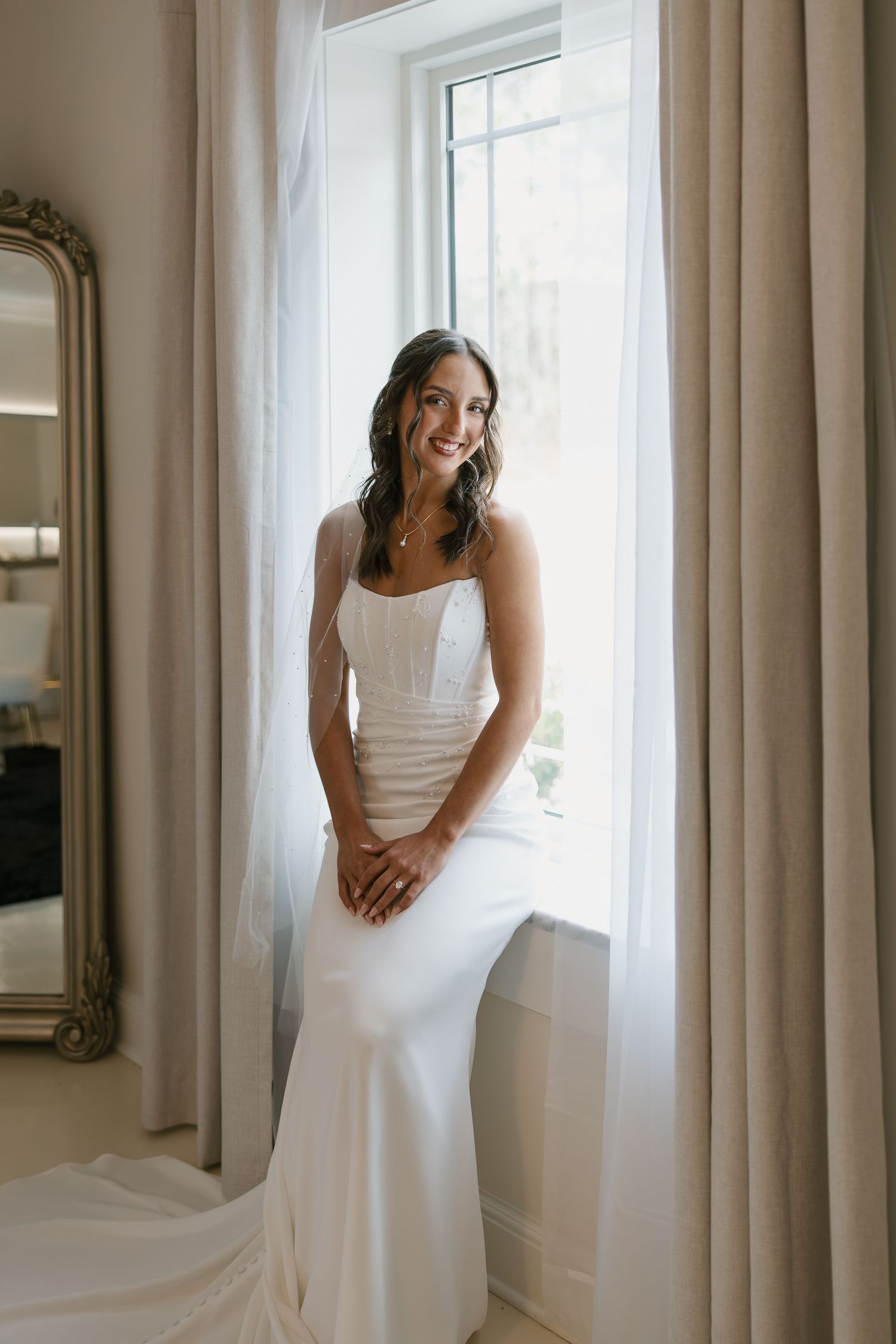 A bride in a white wedding dress stands elegantly by a window with flowing curtains in a bright, airy room.