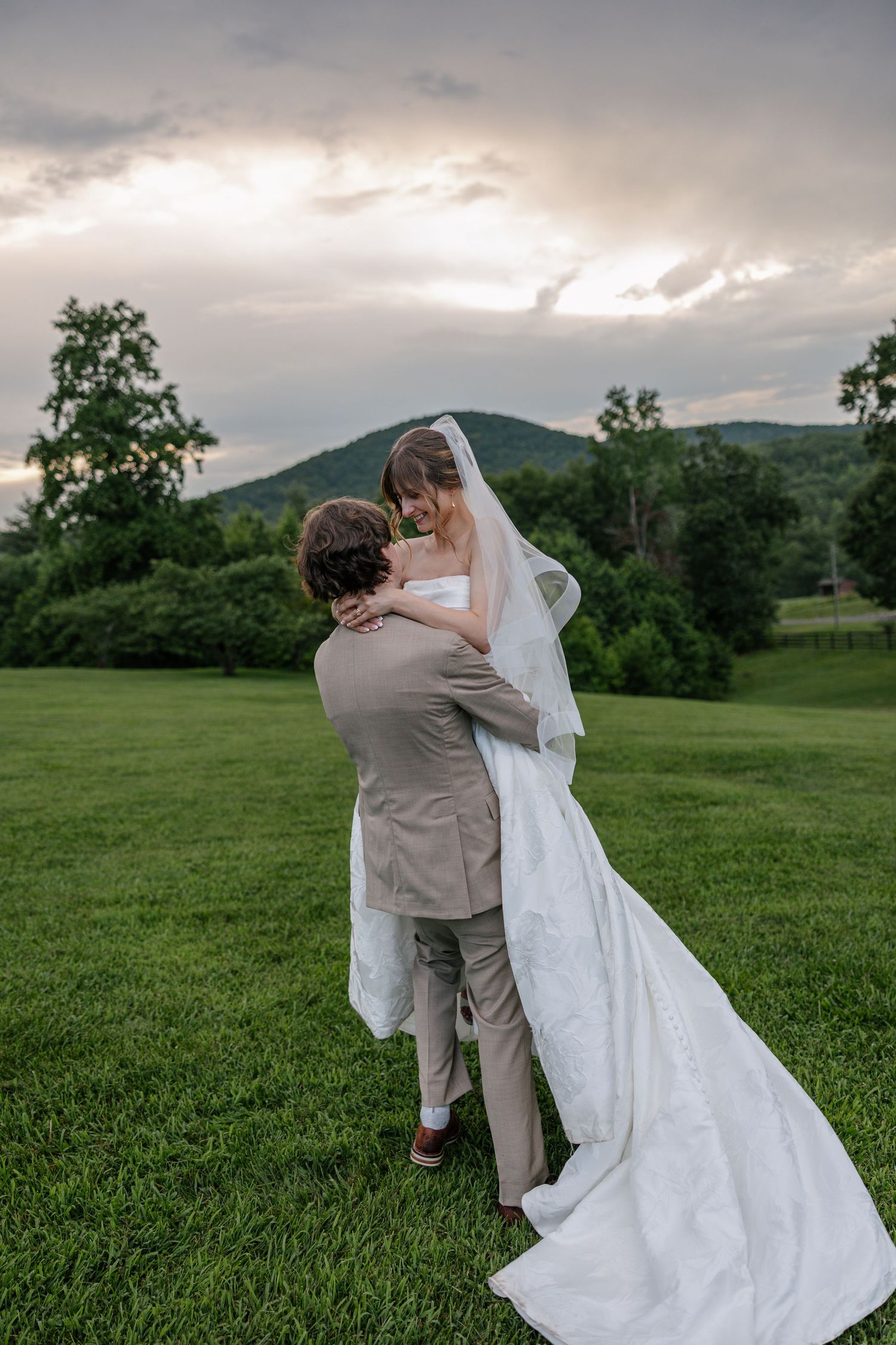 A newlywed couple embraces on a lush green hillside at sunset with mountains and dramatic clouds in the background.
