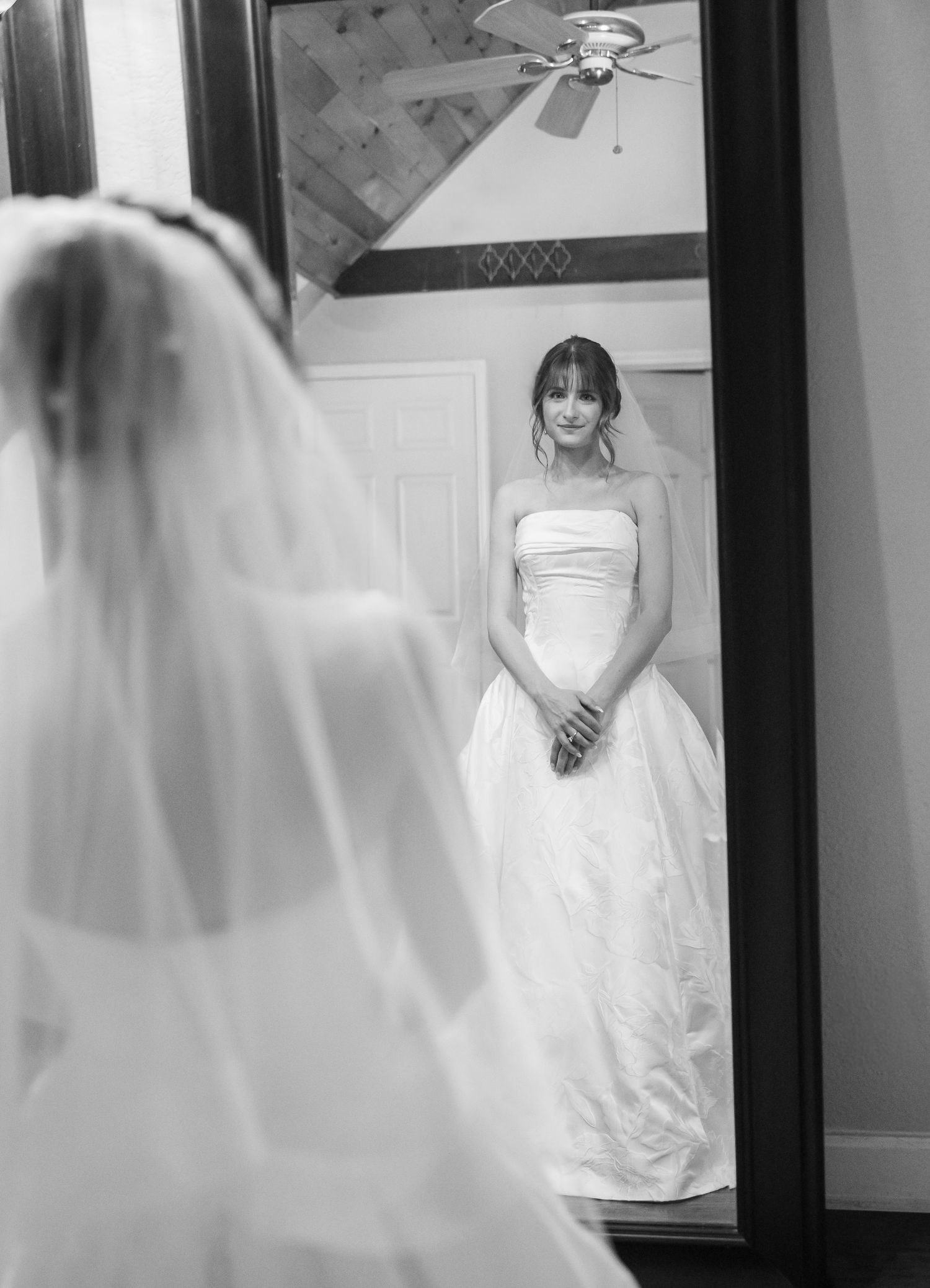A black and white photograph showing a bride in a strapless white gown looking at her reflection in a full-length mirror.
