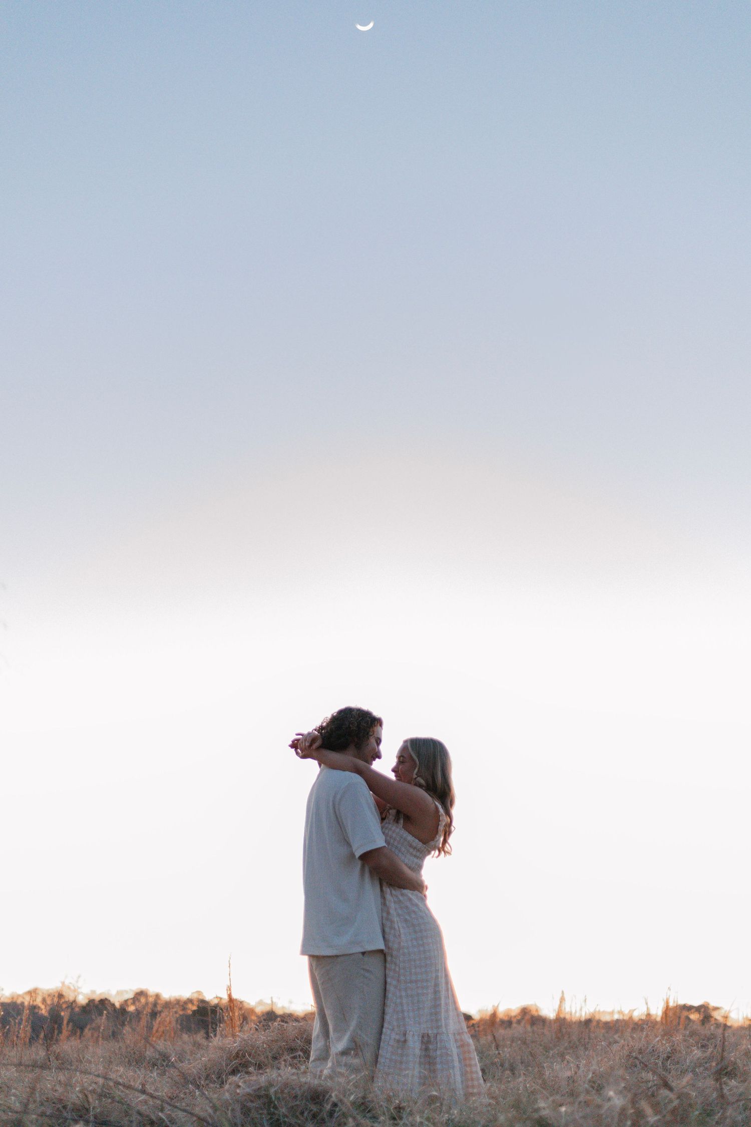 A couple embraces in a field at sunset against a soft pastel sky.