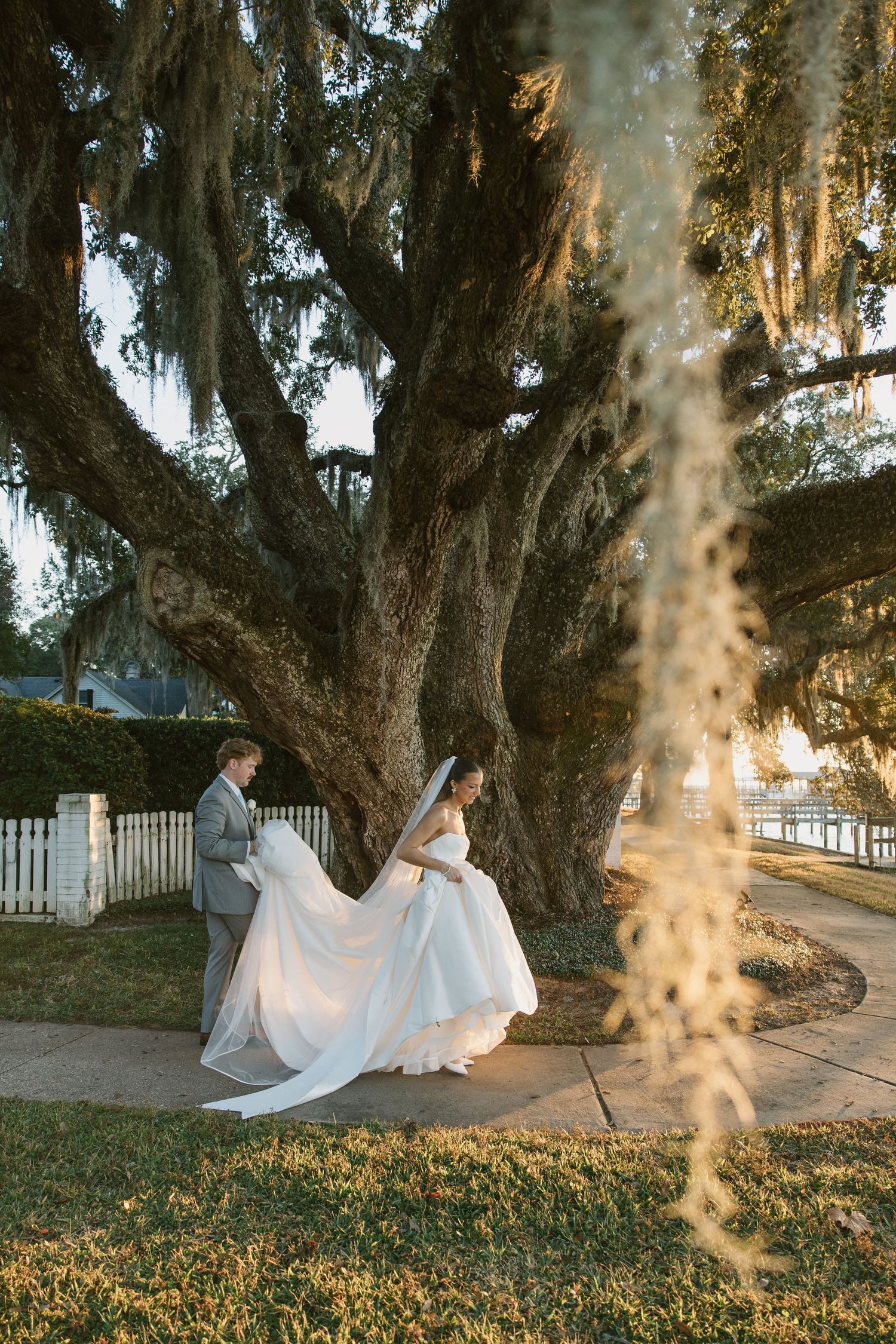 A couple in wedding attire poses under Spanish moss draped trees at sunset in a romantic garden setting.