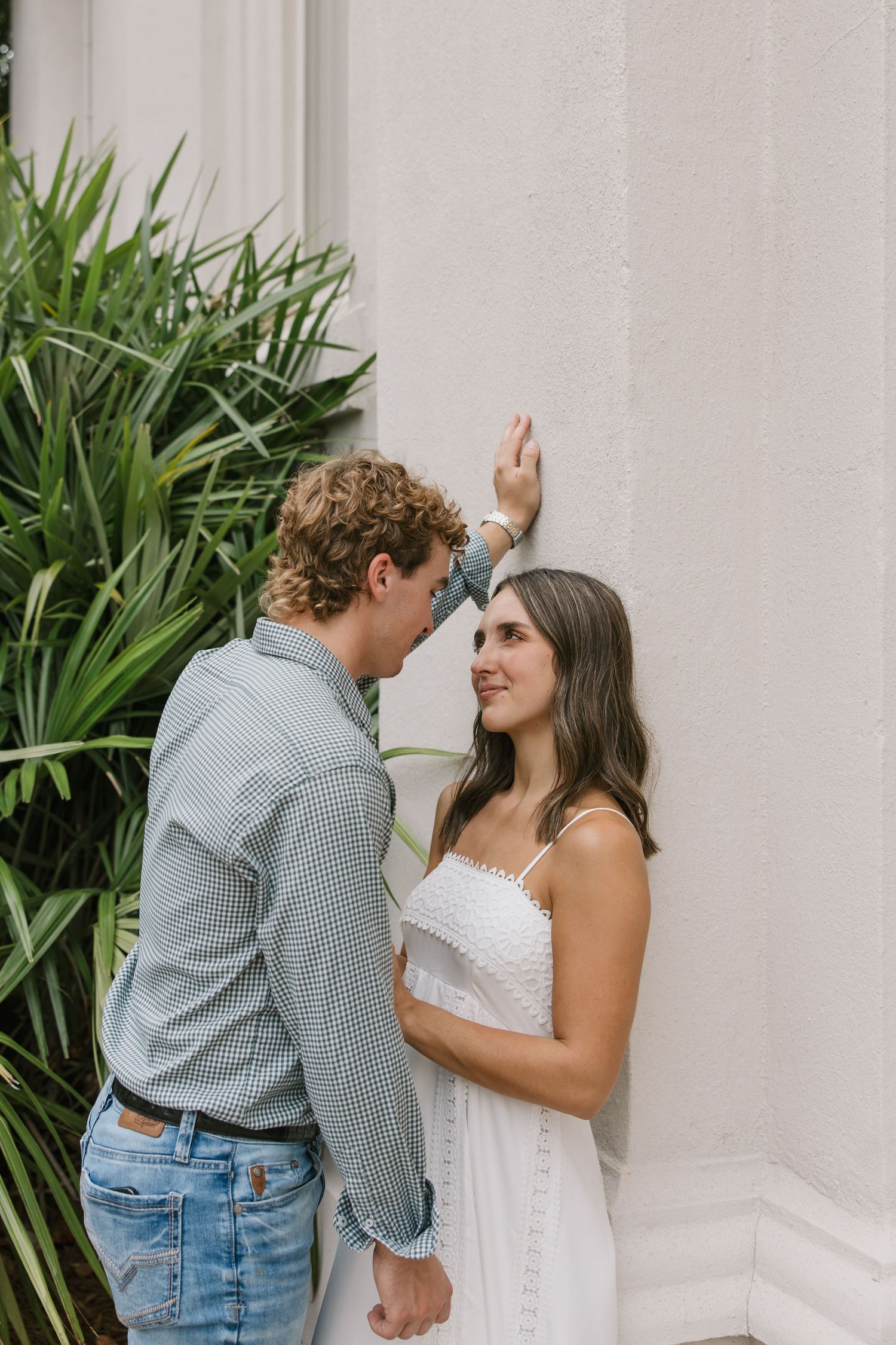 A couple shares a romantic moment against a white wall with tropical plants in the background.