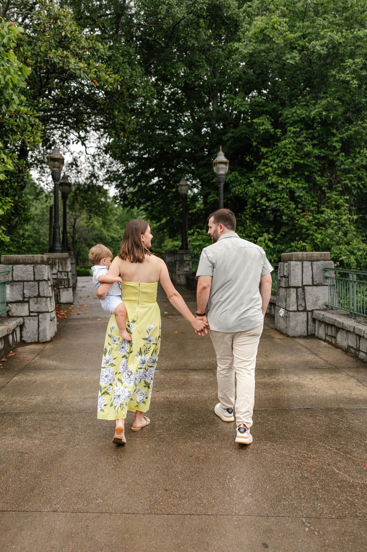 A family walks together hand in hand on a stone bridge path surrounded by lush green trees and lamp posts.