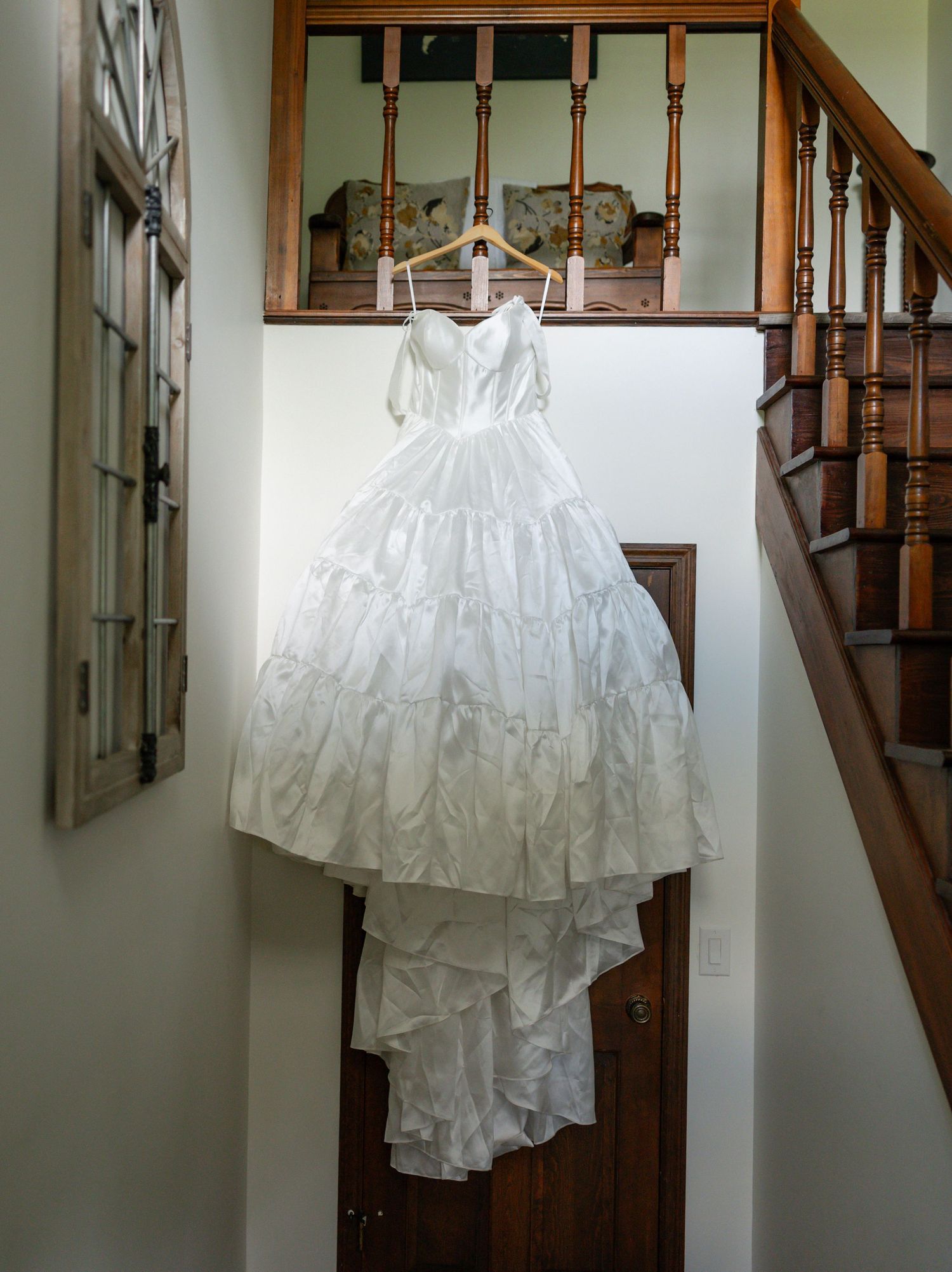 A white wedding dress with layers of ruffled fabric and train hangs elegantly on a hanger against a wall near a wooden staircase.