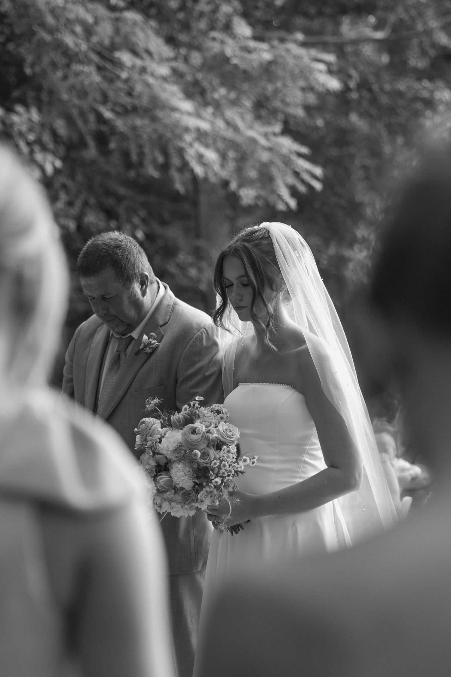 A black and white wedding ceremony moment showing a couple viewing their bouquet in a garden setting.