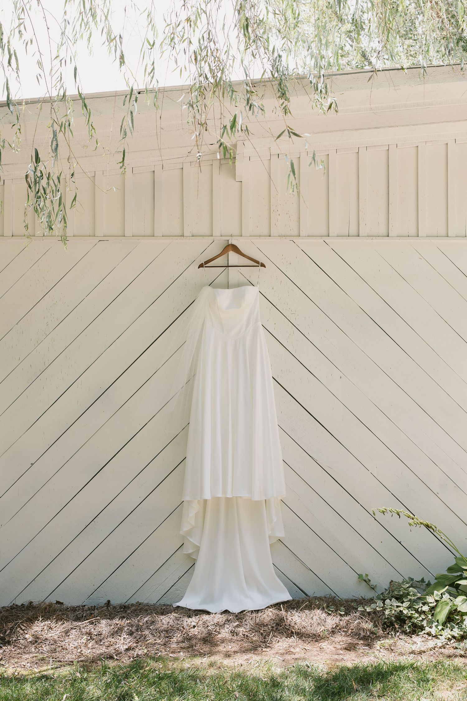A simple white wedding dress hangs on a wooden hanger against a cream-colored diagonal planked fence with willow branches above.