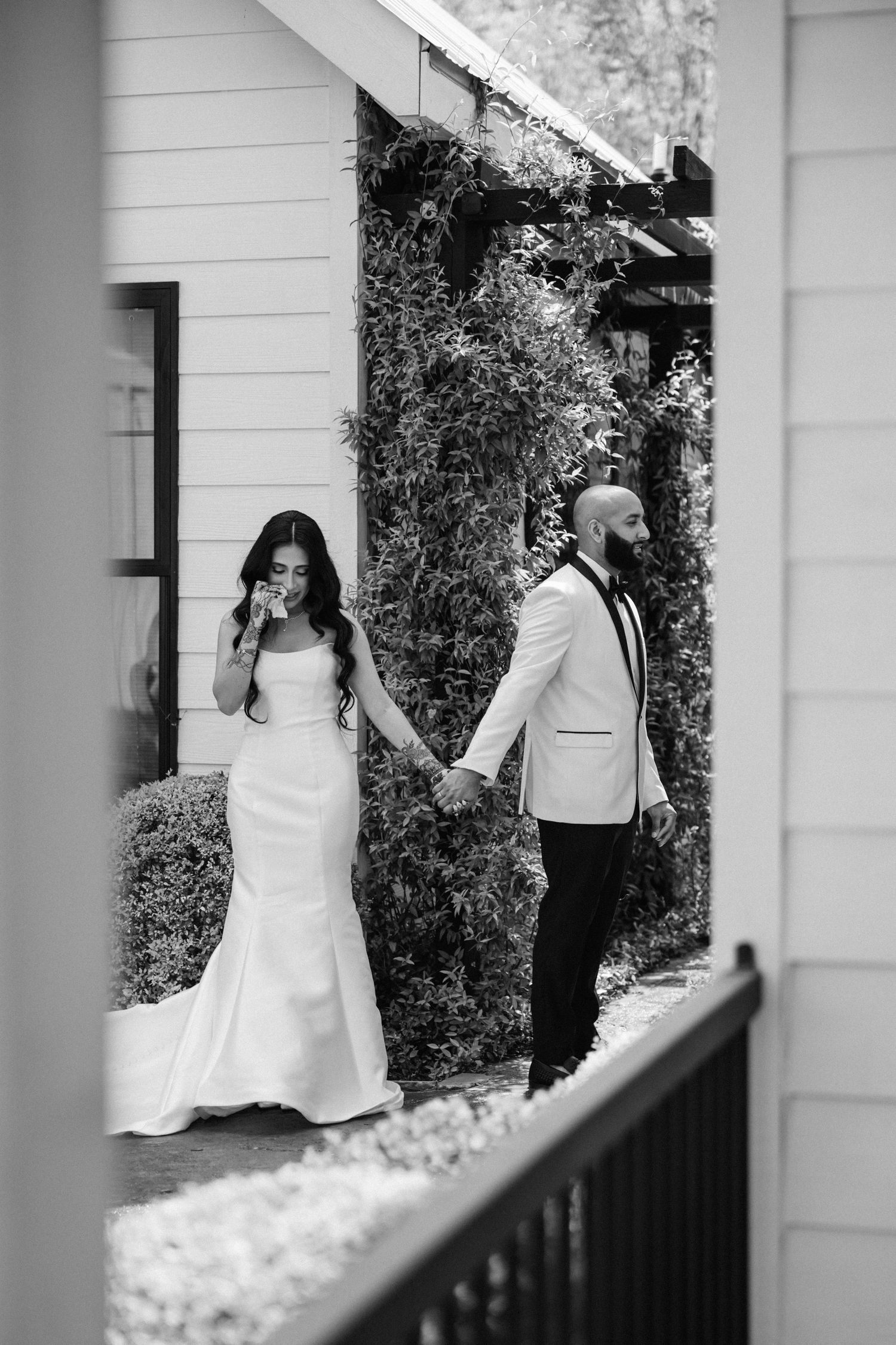 A black and white wedding photo showing a couple holding hands while standing on opposite sides of a building corner.