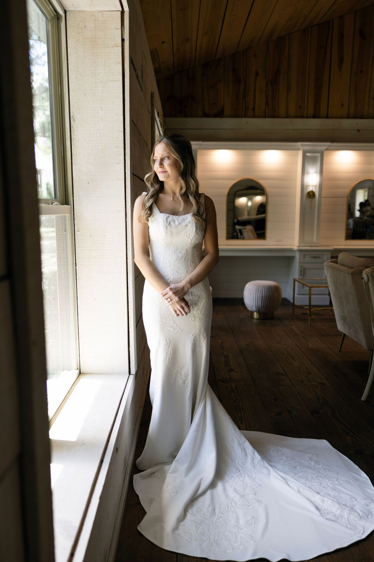 Elegant white wedding gown with lace details and long train shown in rustic wooden interior with natural window lighting.