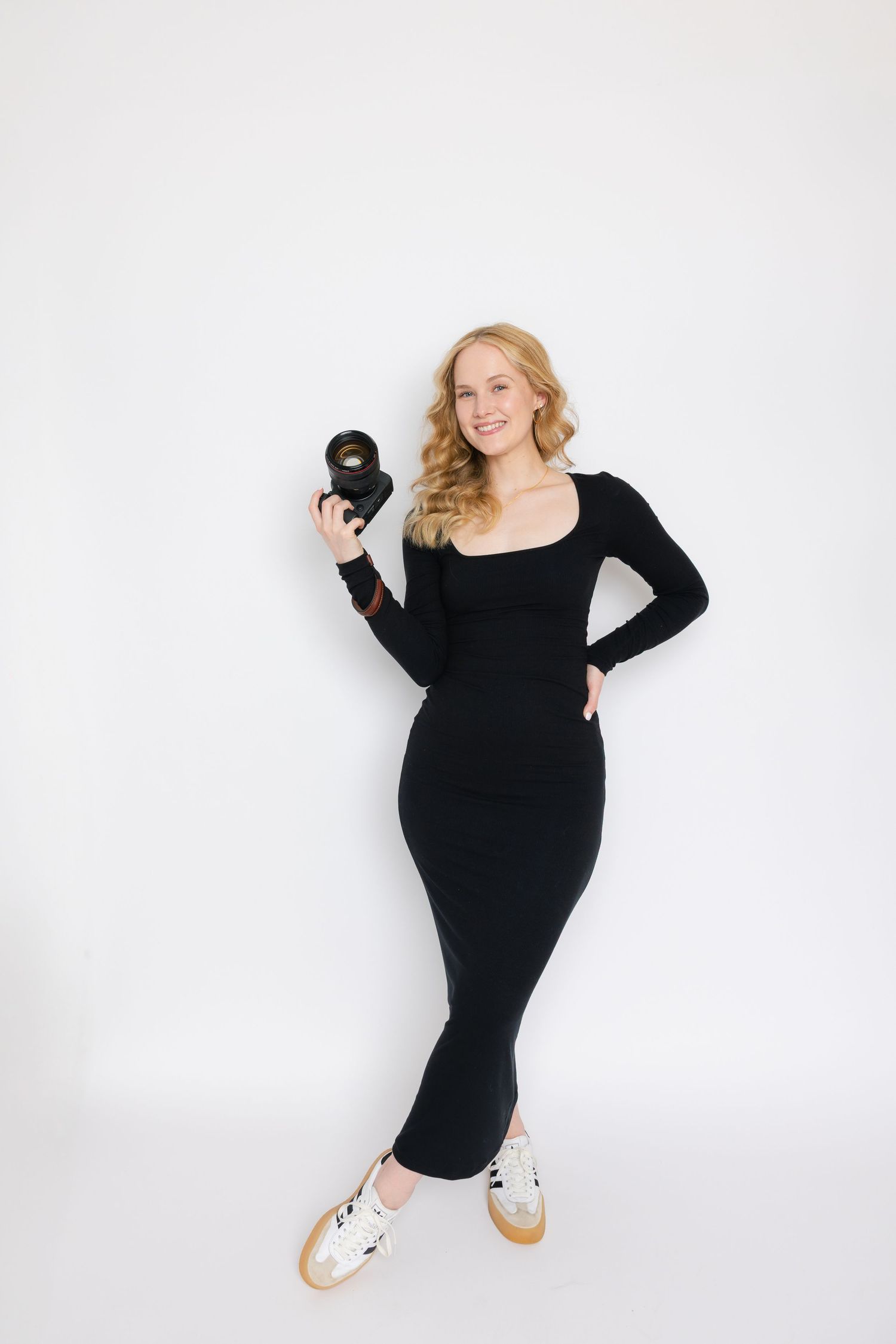 Woman in black bodycon dress holds dumbbells during workout while wearing stylish white sneakers against white backdrop.