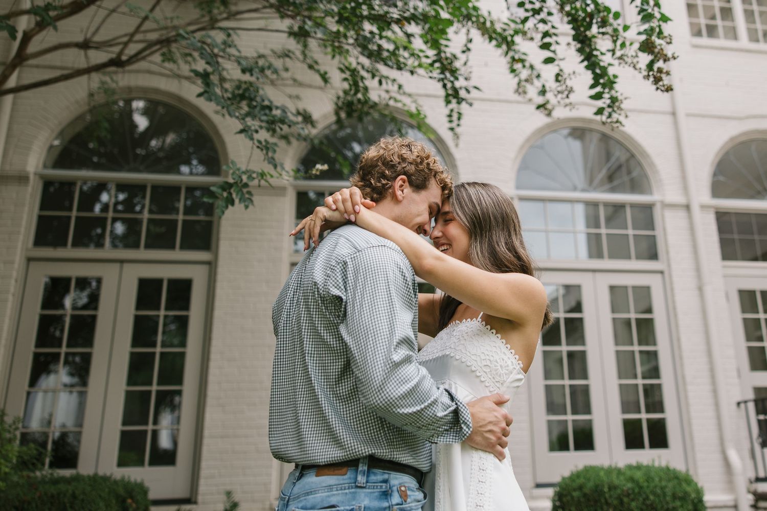 A romantic couple shares an intimate embrace in front of an elegant white building with arched windows and greenery.