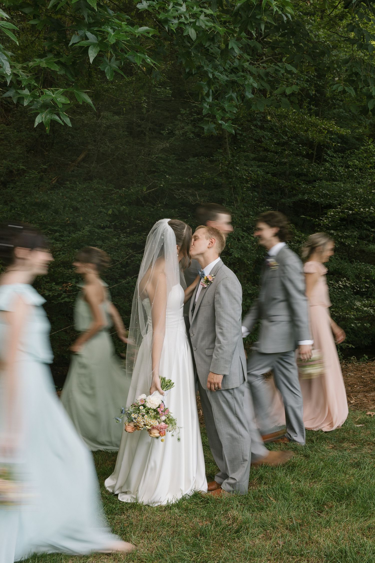 Motion blur captures wedding party walking in a row through green garden setting creating dreamy artistic effect.